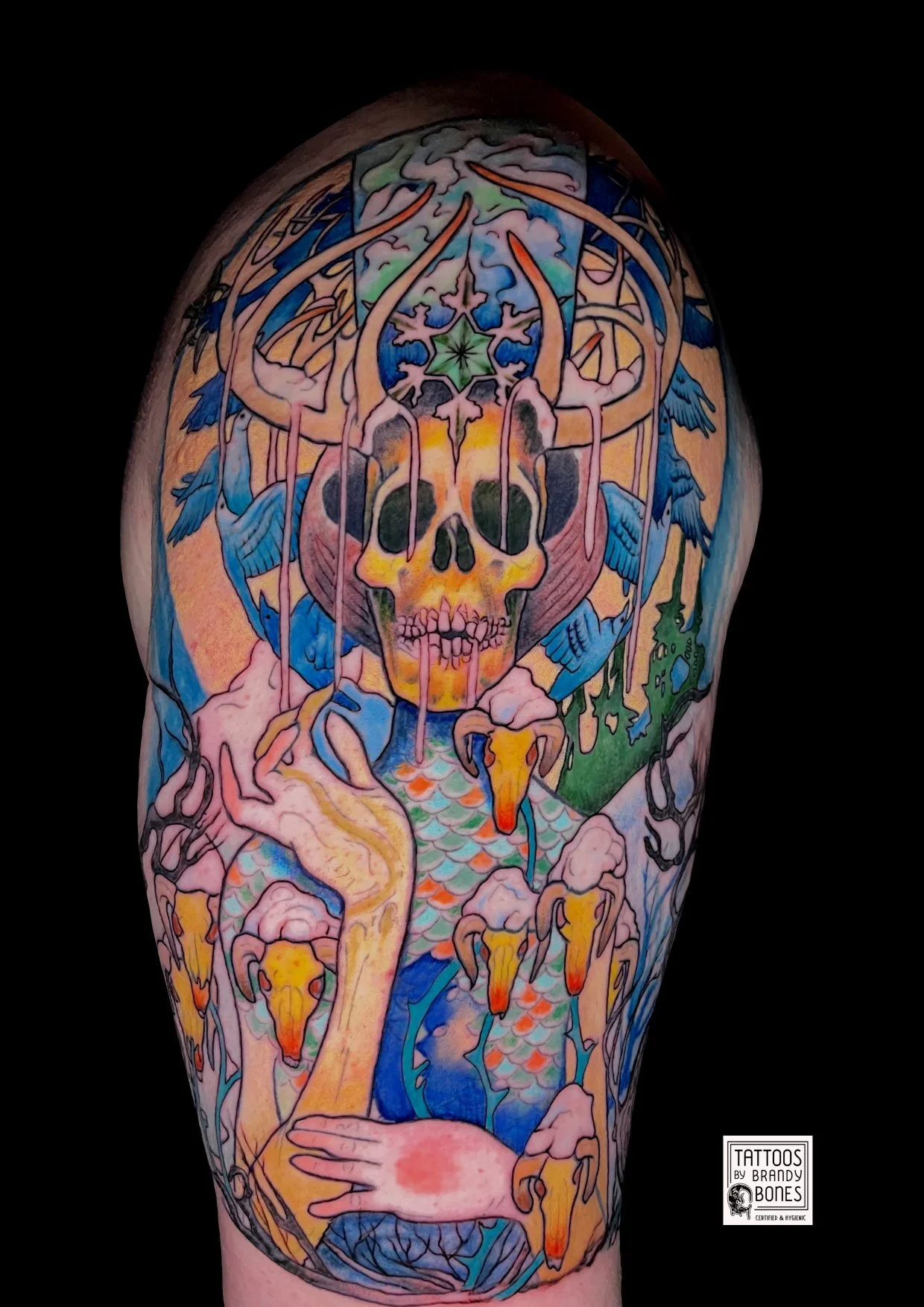 Colorful tattoo of a skull with abstract elements, birds, and a hand on a person's arm.