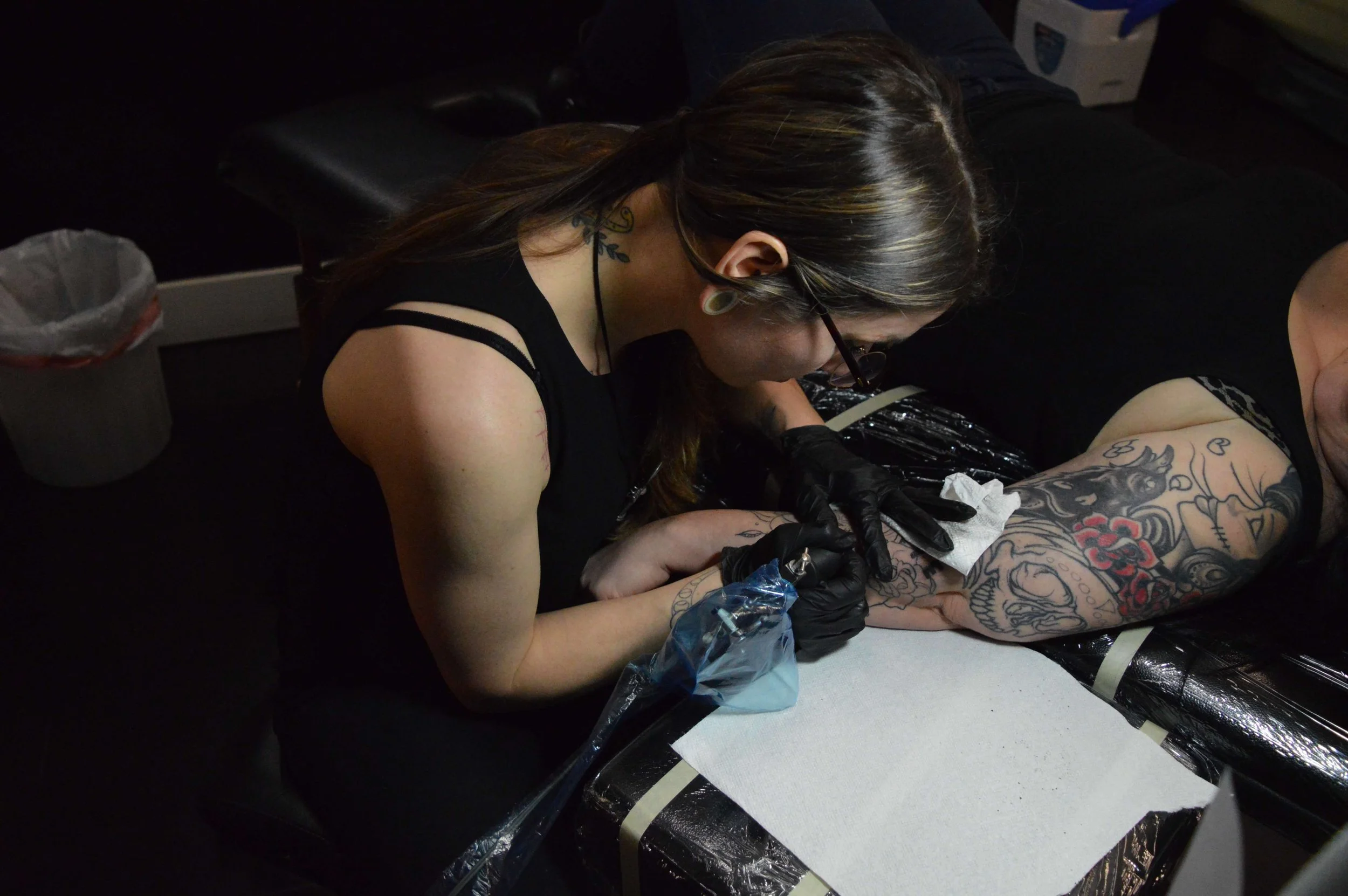 A woman with tattoos in black gloves is tattooing a person's arm with colorful designs, working intensely in a dimly lit environment.