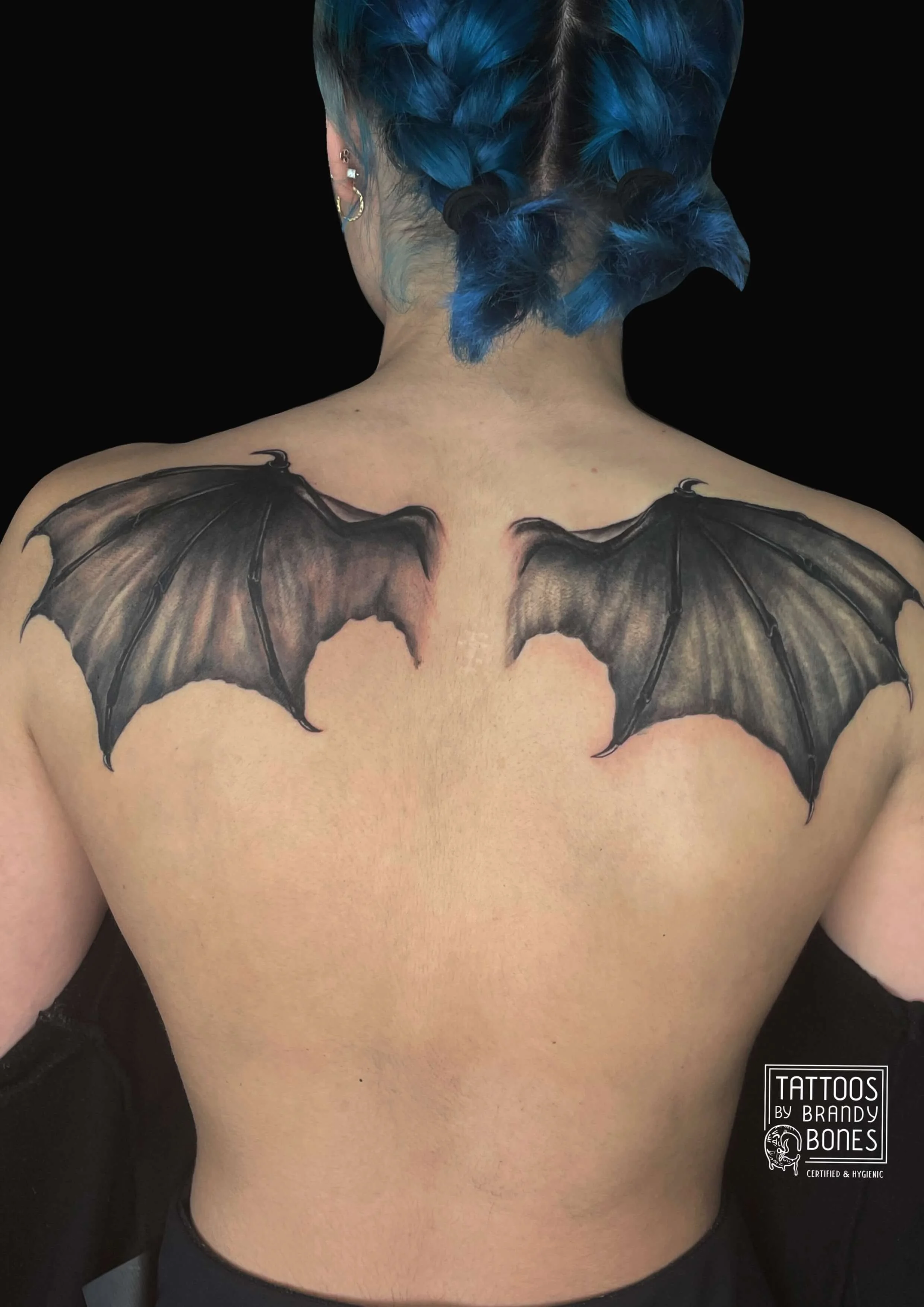 Back view of a person's upper body with bat wings tattoo on the shoulders, with blue braided hair and multiple earrings.