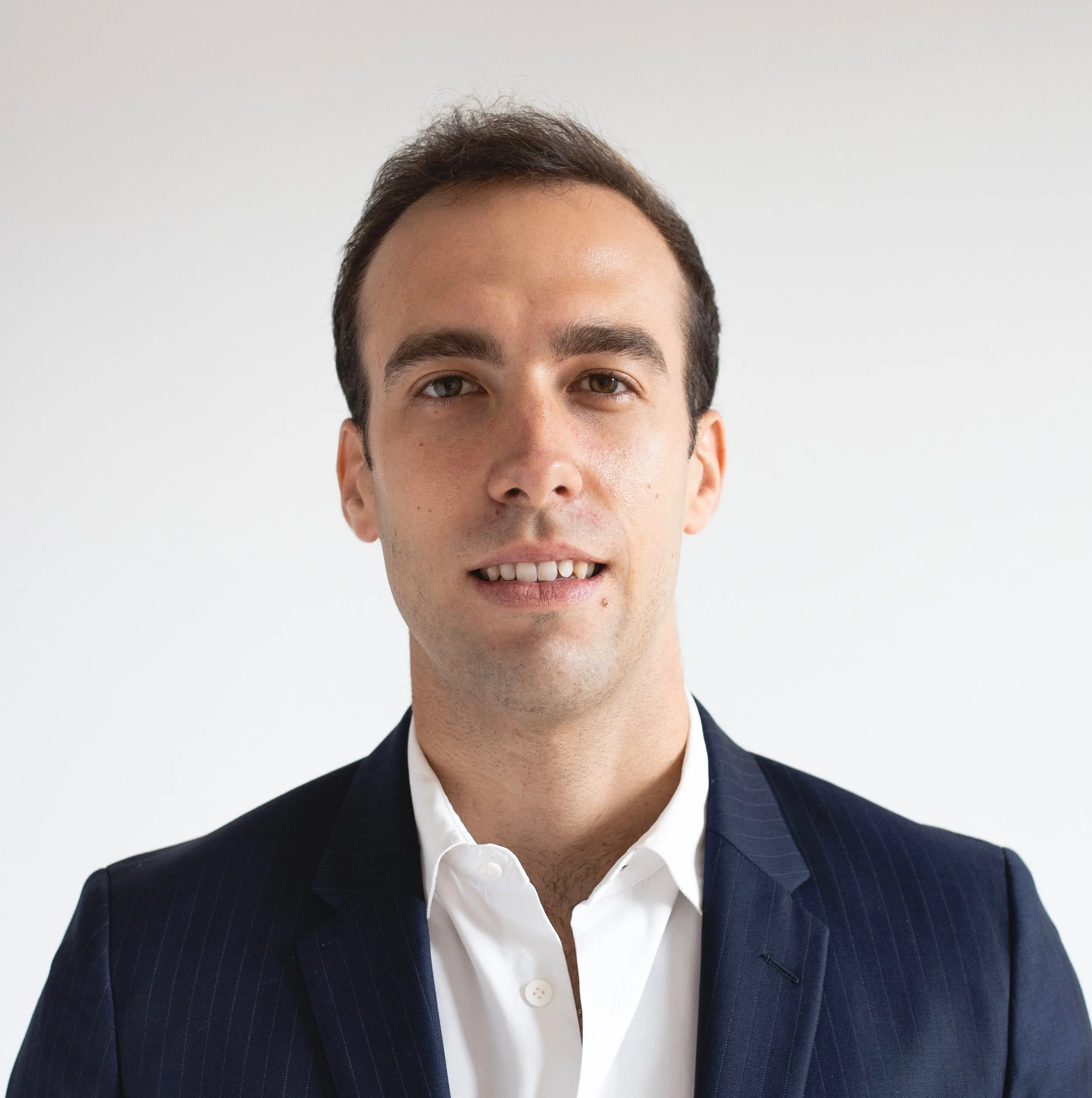 Portrait d'un homme avec des cheveux bruns courts, portant un costume noir à fines rayures et une chemise blanche, souriant légèrement, contre un fond blanc.