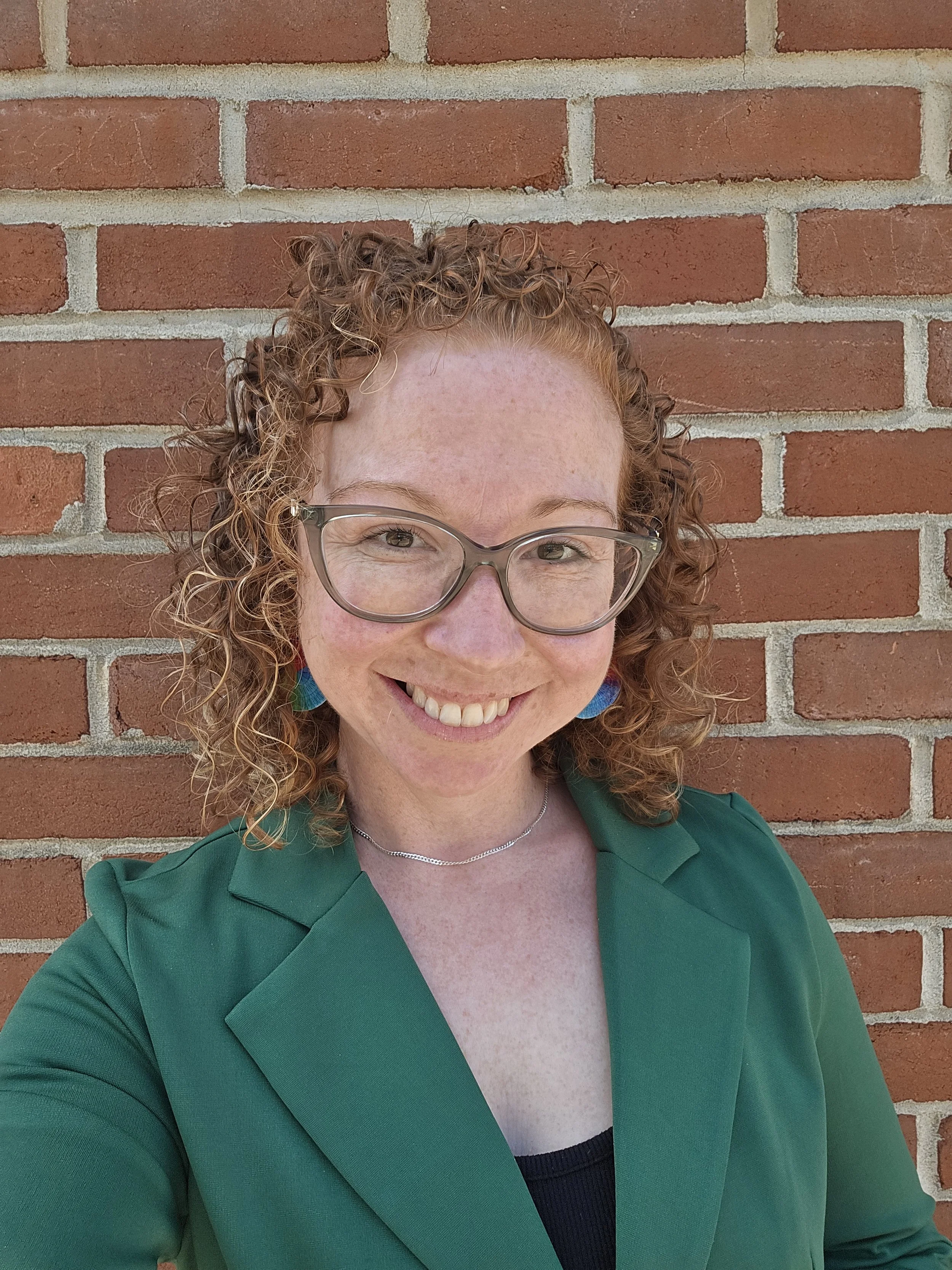 A woman with curly red hair and glasses smiling in front of a brick wall, wearing a green blazer and a black top.