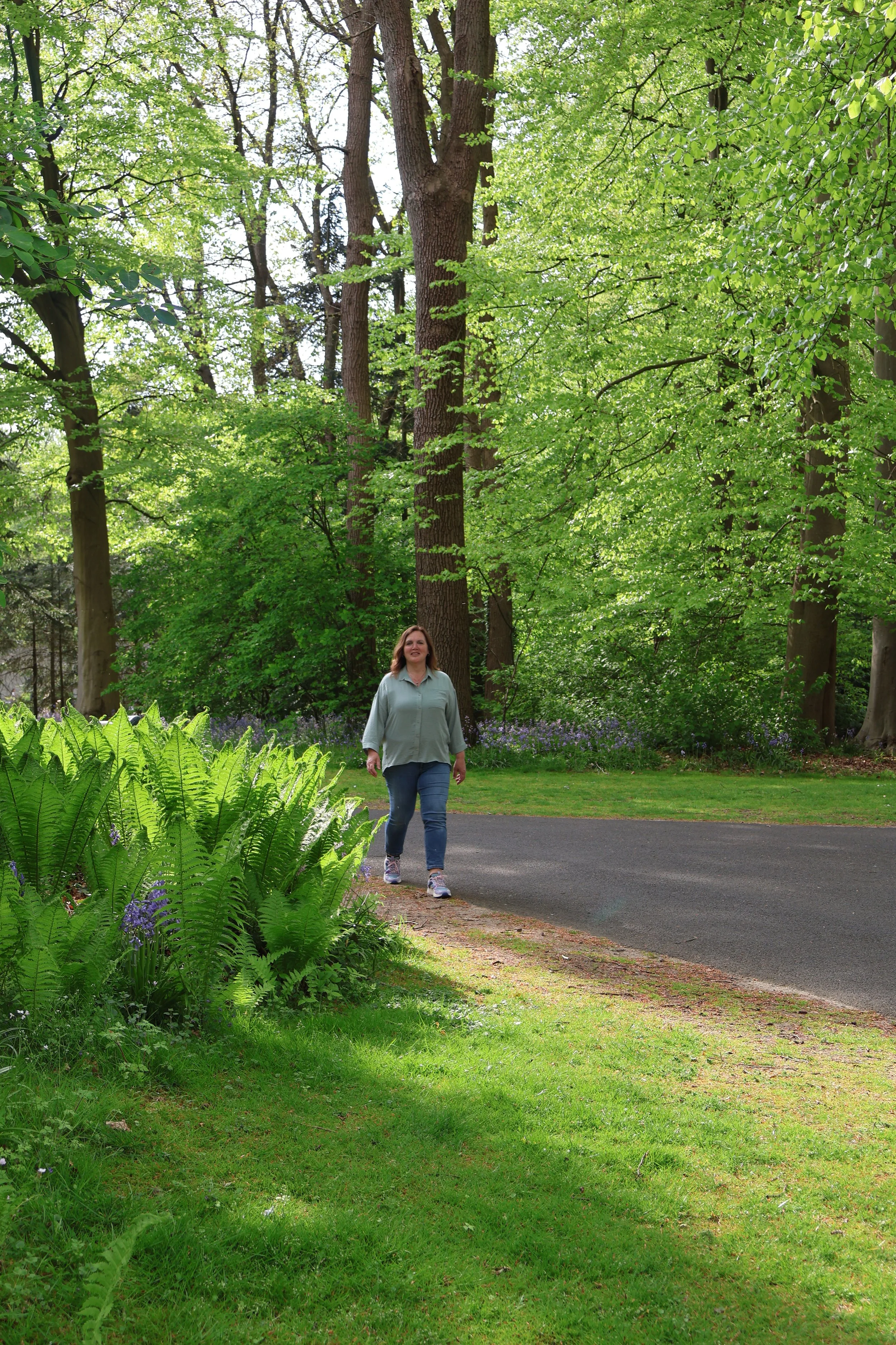 Vrouw loopt langs een groen gazon en bloemen, omgeven door grote groene bomen in een park op een zonnige dag.
