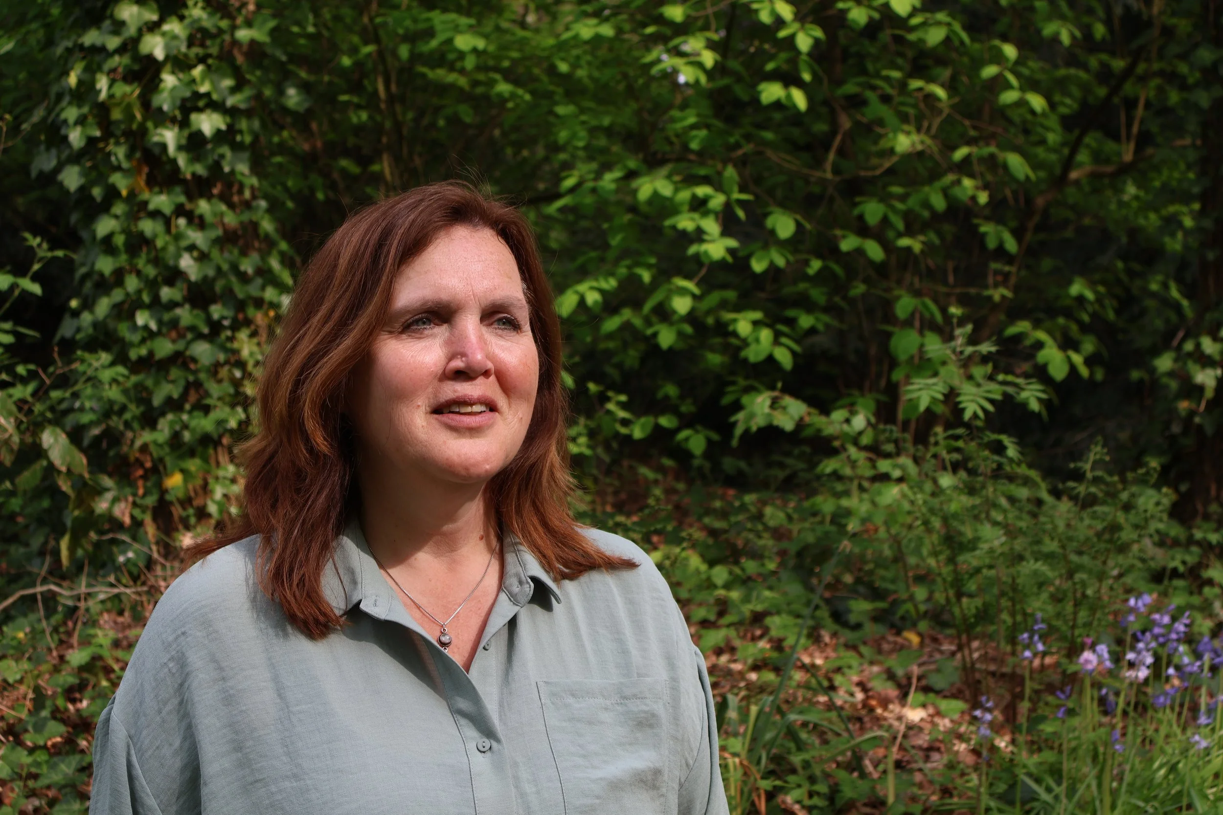Een vrouw met bruin haar die naar links kijkt, staand voor een groene, bosrijke achtergrond met bladeren en bloemen.
