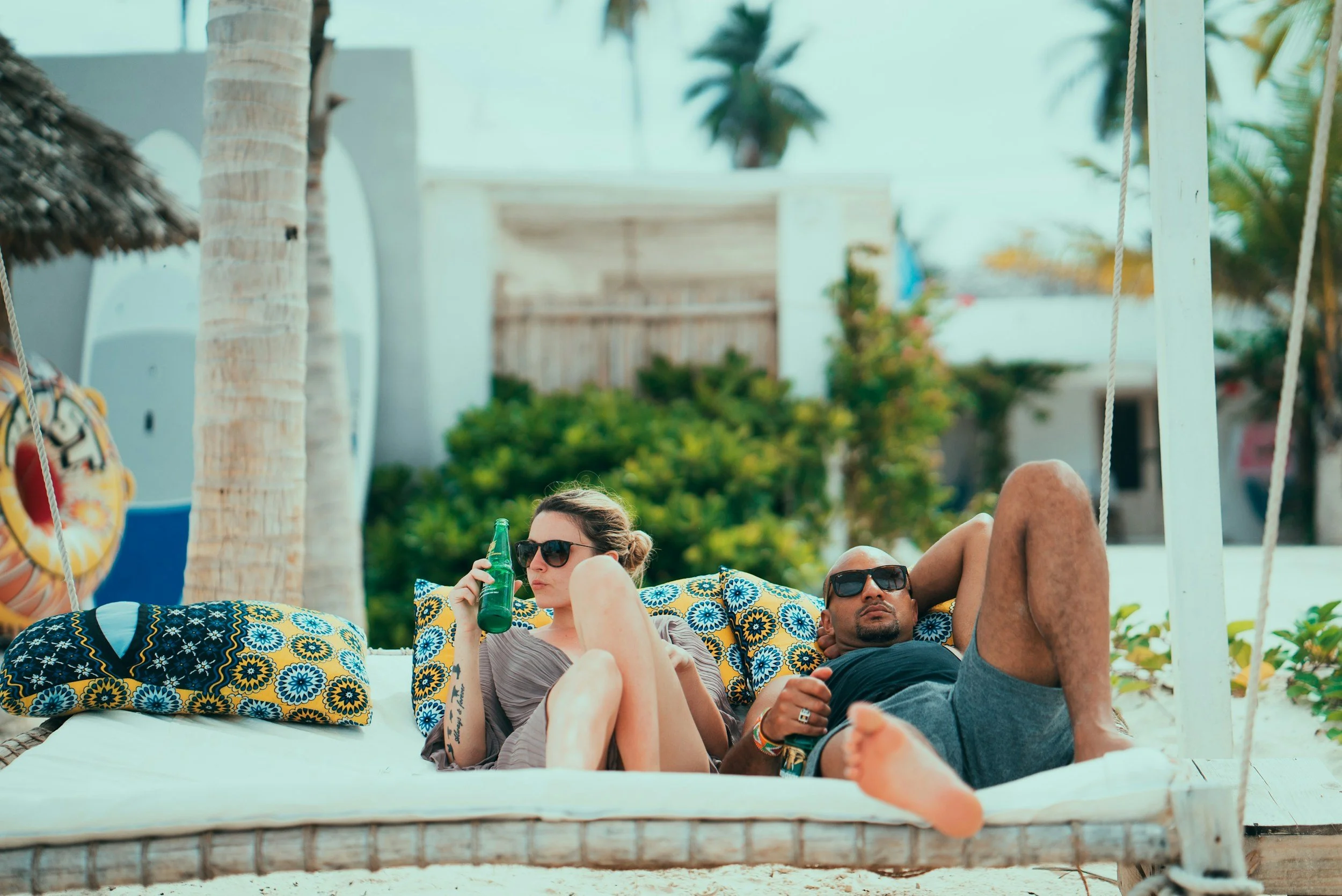 A man and woman relax on a beach swing bed with tropical pillows, surrounded by palm trees, holding drinks, under sunny weather.