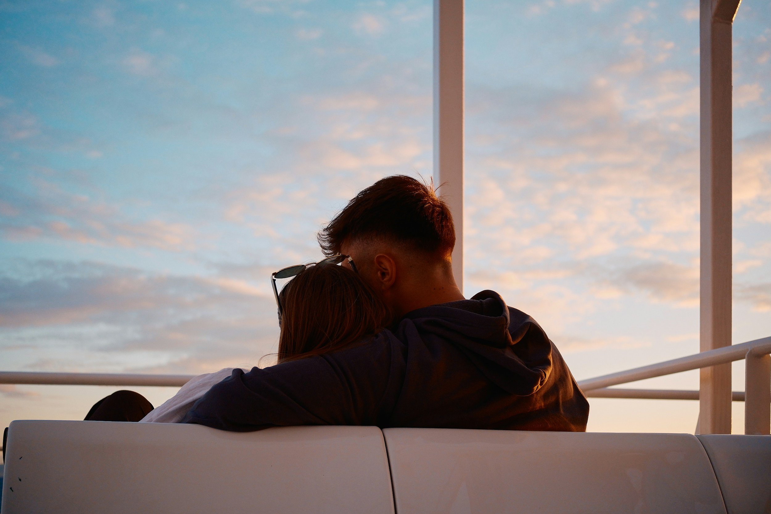 A couple sitting on a white bench embracing, with a sunset sky and clouds in the background.