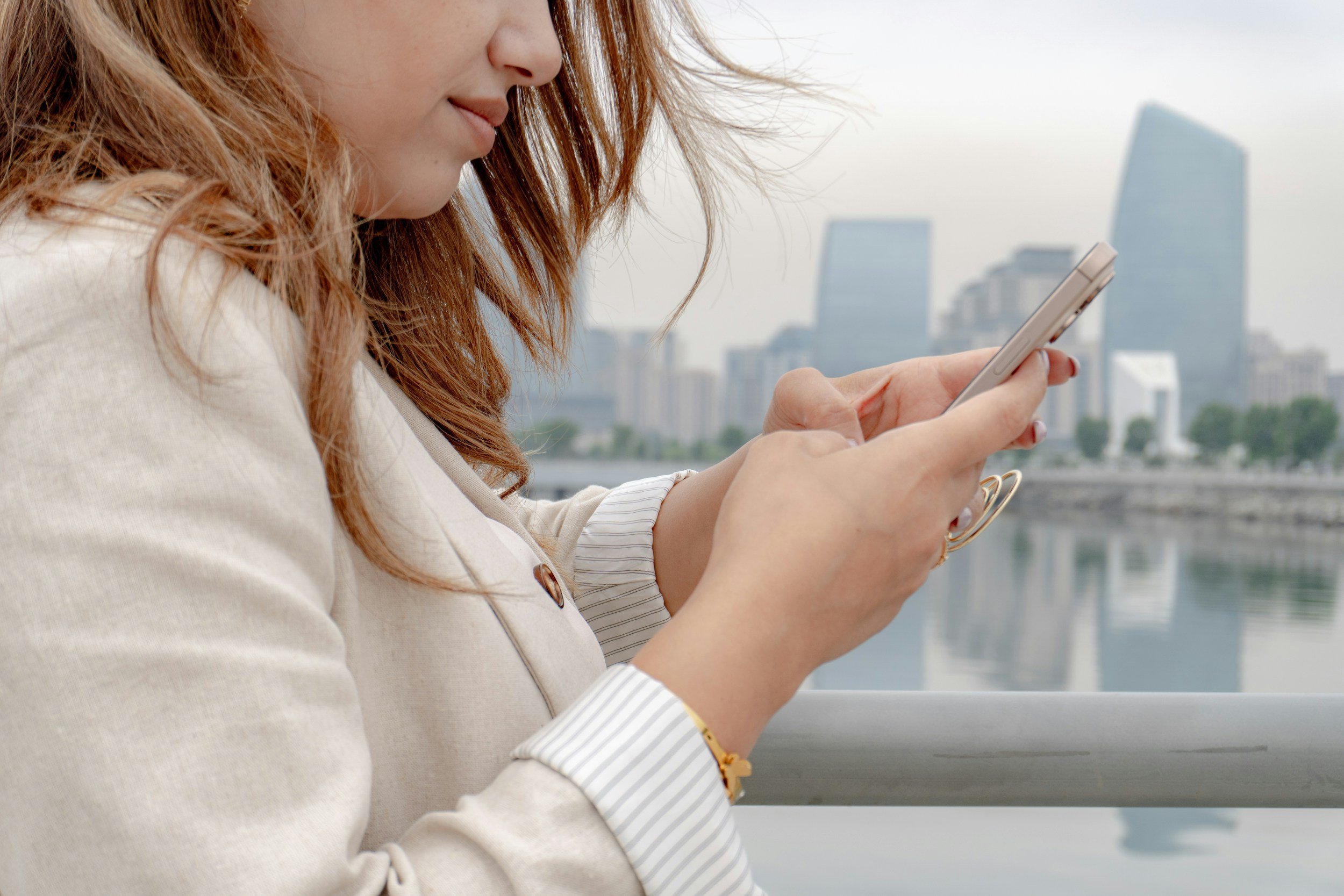 Woman with red hair dressed in a beige blazer using a smartphone with city buildings in the background.