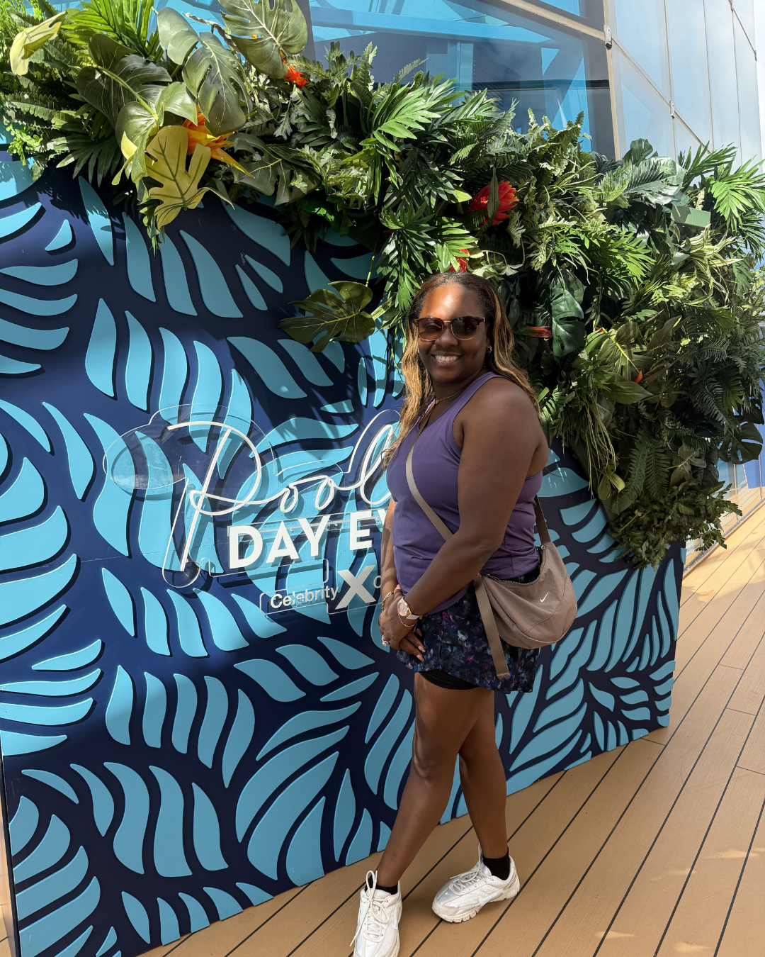 A woman standing in front of a tropical-themed backdrop with lush green plants and a blue patterned wall, smiling at the camera. She is wearing sunglasses, a purple tank top, black shorts, and white sneakers, with a beige crossbody bag.