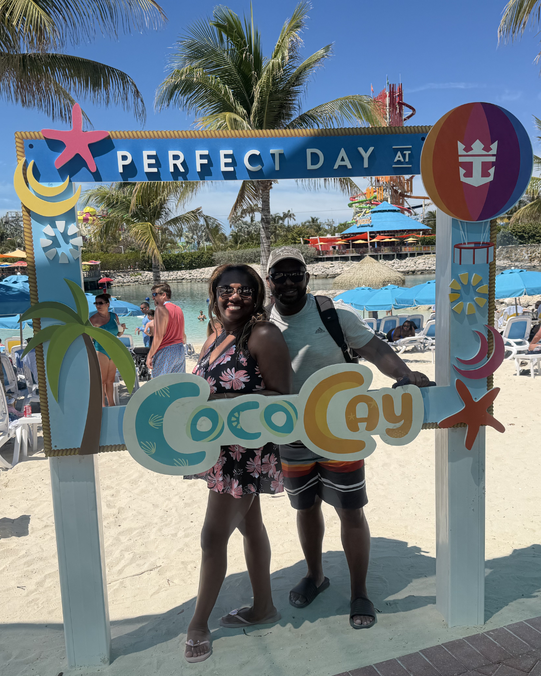 A smiling couple at Coco Cay, Bahamas, standing inside a colorful beach-themed photo frame with the words 'Perfect Day at Coco Cay'. They are at the beach with umbrellas, lounge chairs, and palm trees in the background.