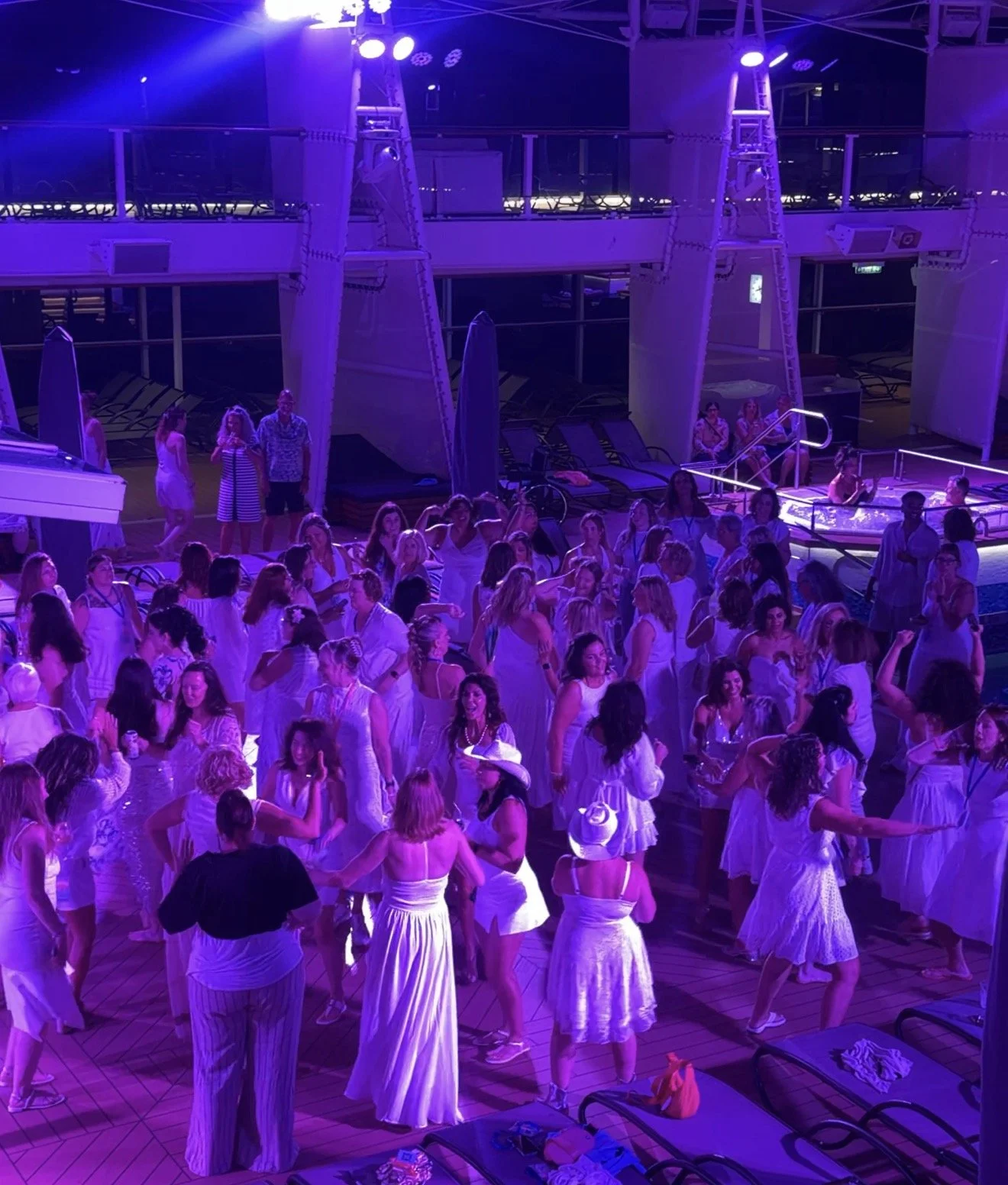 Group of women dancing and socializing on a cruise ship deck illuminated with purple lighting, with lounge chairs and a hot tub in the background.