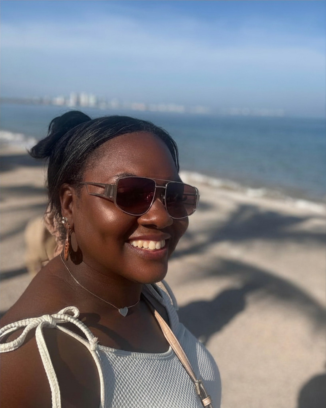 A woman smiling on a beach, wearing sunglasses, earrings, a necklace, and a white outfit, with the ocean and distant city skyline in the background.