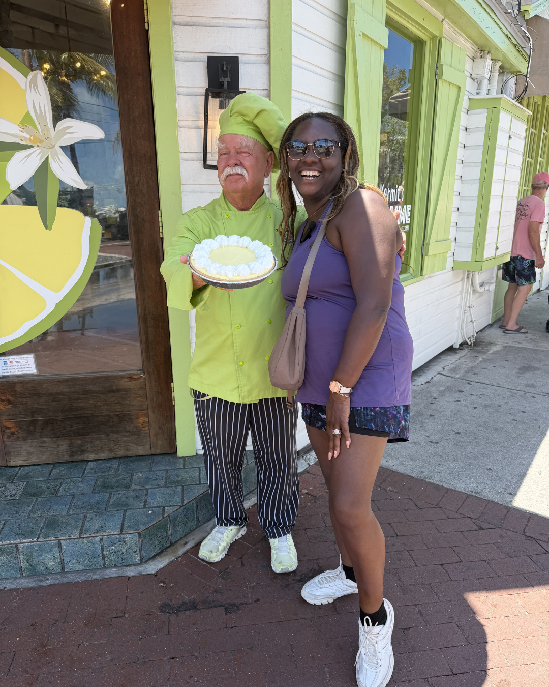 A woman in sunglasses smiling, standing beside a chef holding a small cake outside a restaurant with green trim. The chef is wearing a green uniform and hat, and striped pants. The restaurant has painted white wooden siding and green shutters, with outdoor seating visible.