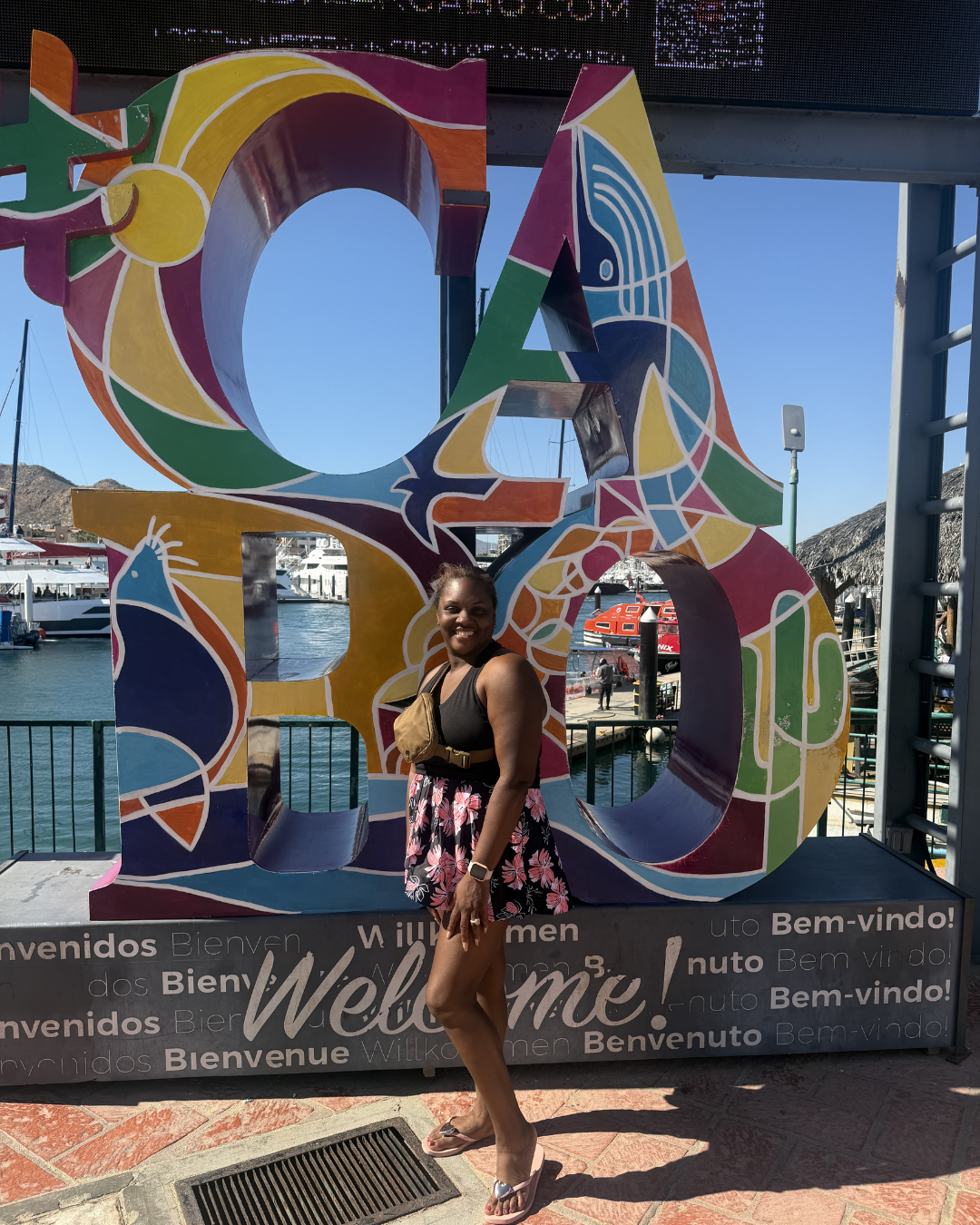 A smiling woman standing in front of a colorful 'LOVE' sign at a marina, with boats and clear blue sky in the background.