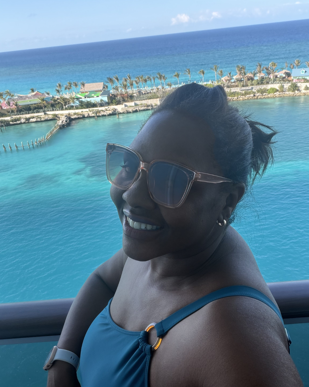 A woman smiling with sunglasses on, enjoying a view of the ocean and coastline with houses and palm trees in the background.