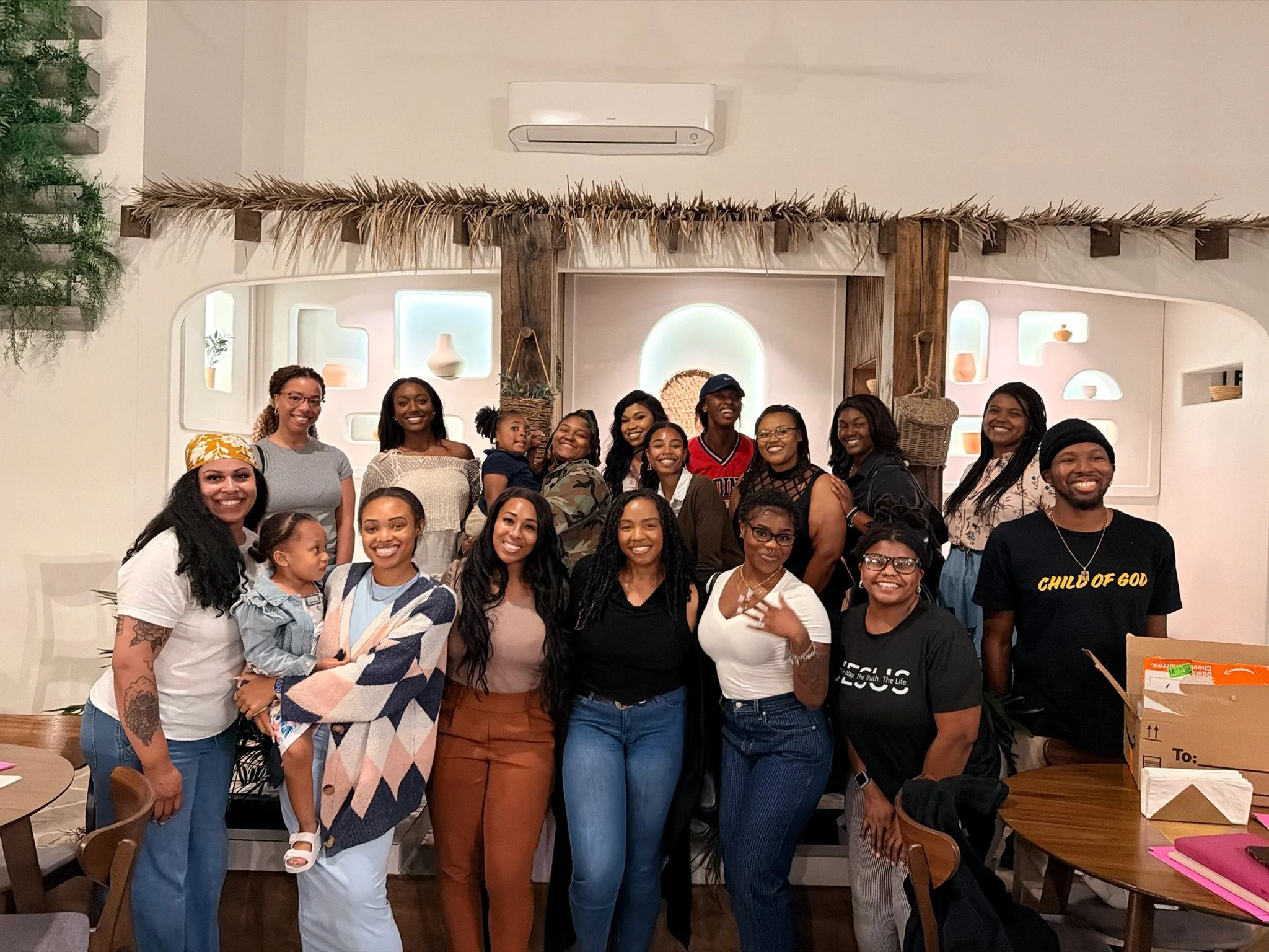 A group of women and children smiling and posing together in a restaurant or cafe. The women are dressed casually, and some are wearing glasses. There are tables, chairs, and a decorative wall with niches in the background. The setting has a warm and friendly atmosphere.