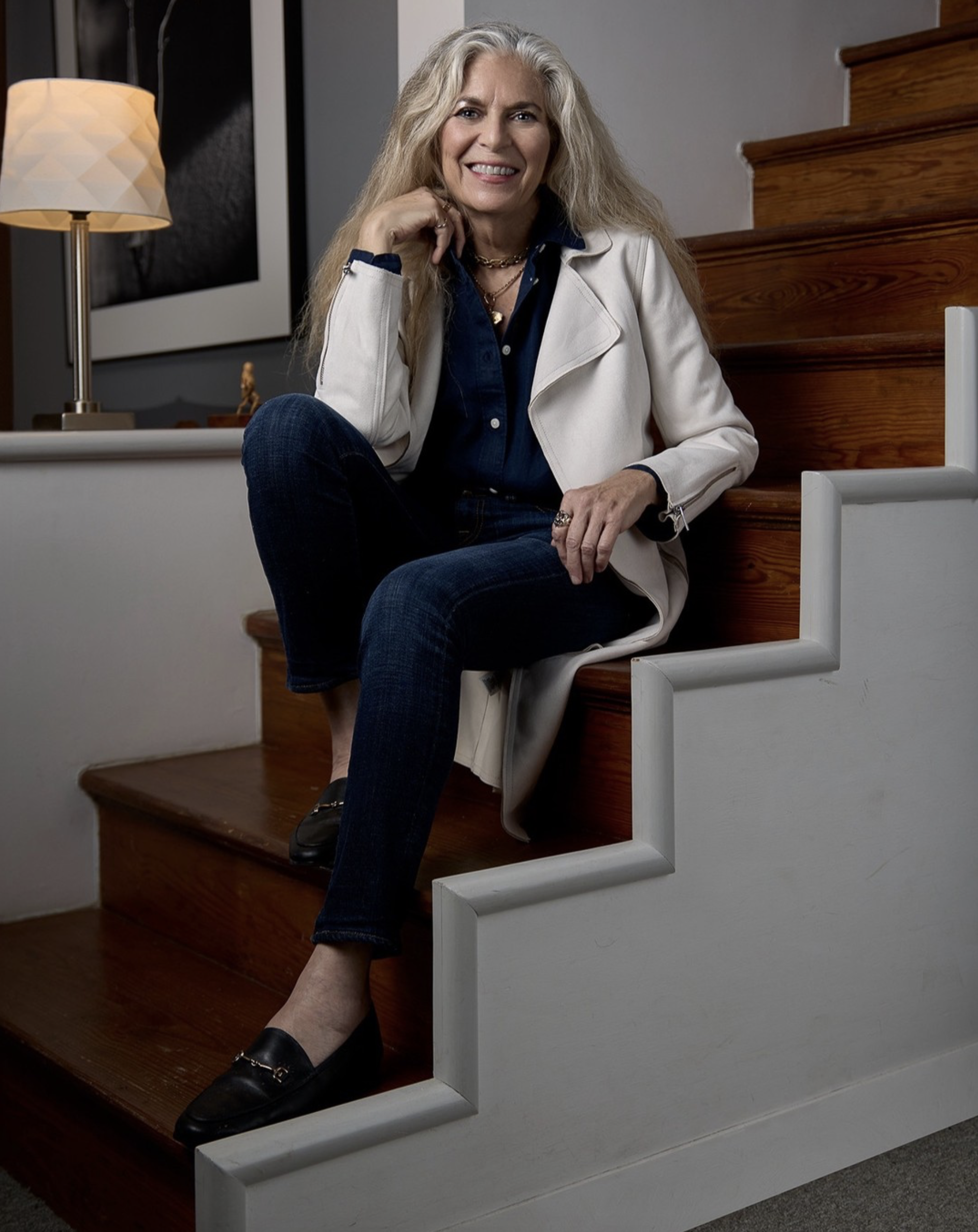 Susan W. Morgan with long blonde hair smiling at the camera, seated on wooden stairs, wearing a white blazer, dark blue shirt, and dark jeans, with a lamp and framed artwork in the background.