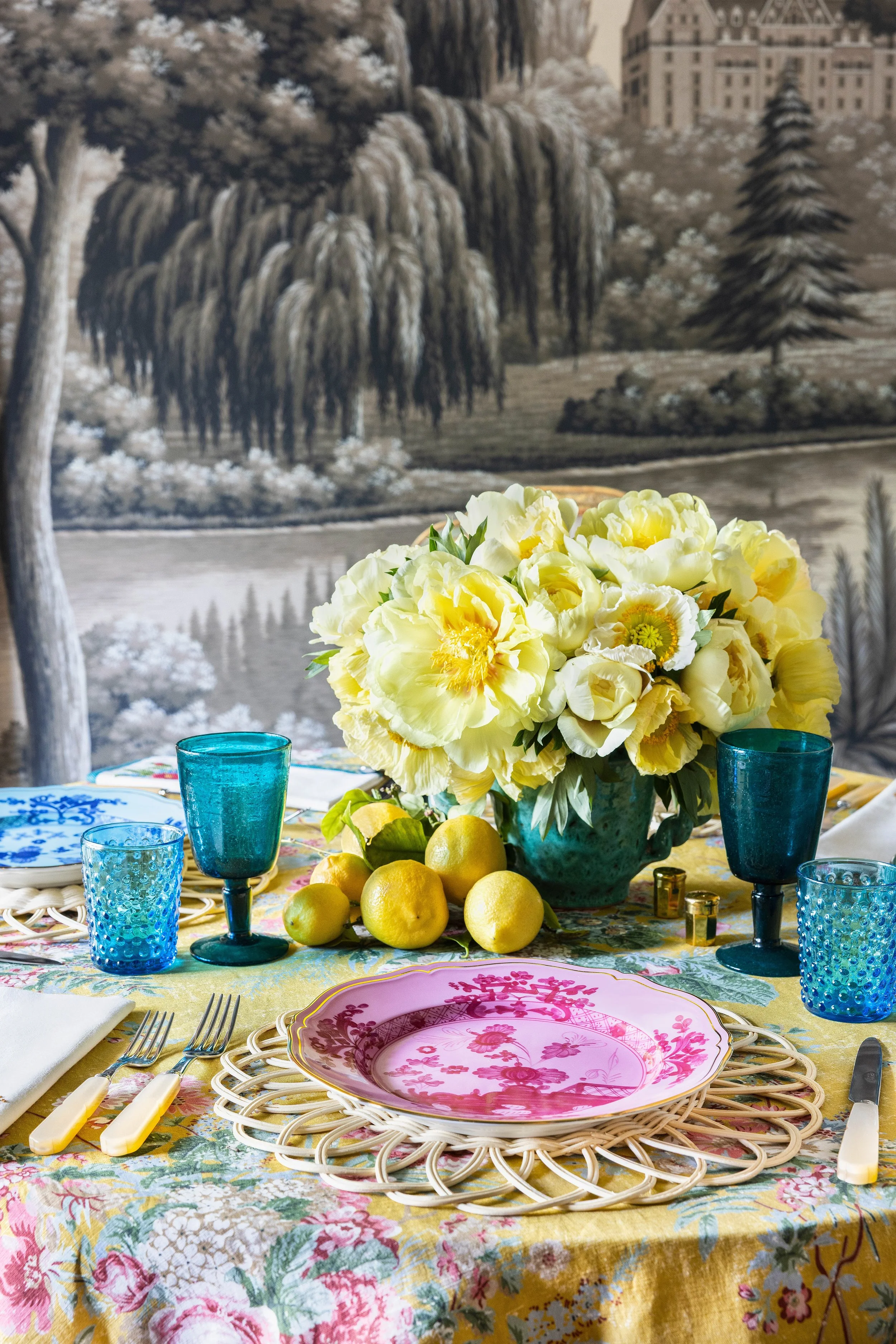 A colorful table setting with pink floral plates, yellow and green lemons, and a large bouquet of white and yellow flowers as a centerpiece. There are four blue textured glasses, a fork and knife with yellow handles, and a yellow tablecloth with a floral pattern. In the background, a black-and-white mural depicts a park scene with trees, water, and buildings.