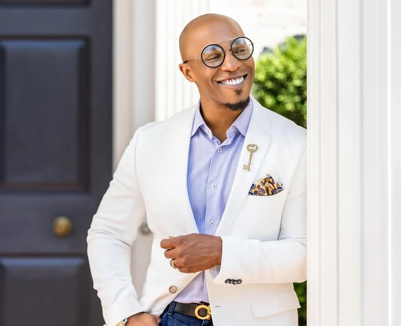 Smiling Cody Damen Jenkins with glasses in a white suit standing at the door of a house.