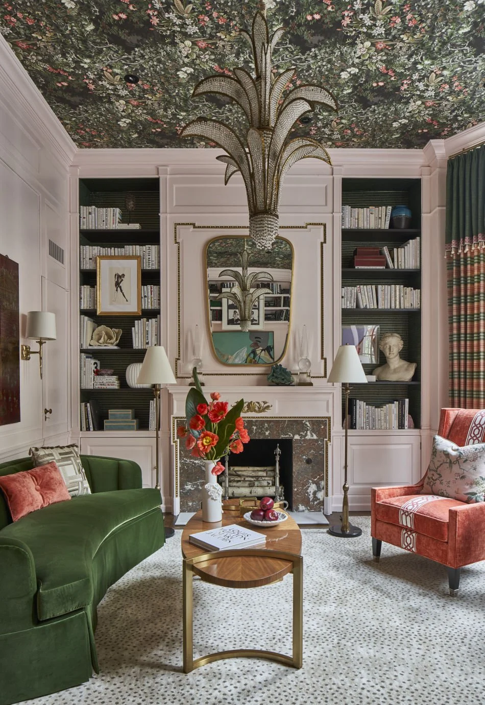 Elegant living room with a green velvet sofa, coral armchair, and a mix of decorative pillows. Central wooden coffee table with a floral arrangement in a white vase, apples, and a book. Fireplace with a marble surround and built-in bookshelves filled with books. Large mirror above the fireplace reflects the room and ceiling. Floral patterned ceiling with a large, ornate chandelier resembling a palm leaf. Wall sconces, a bust sculpture, and a painting add artistic touches. Green curtains frame the windows, and a textured, dotted rug covers the floor.