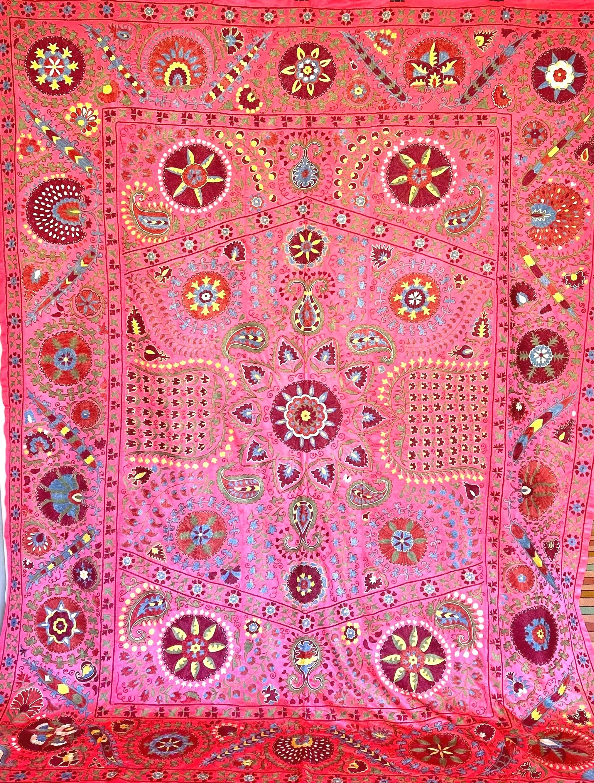A vibrant pink textile with intricate floral and paisley patterns, detailed with embroidery and colorful accents.