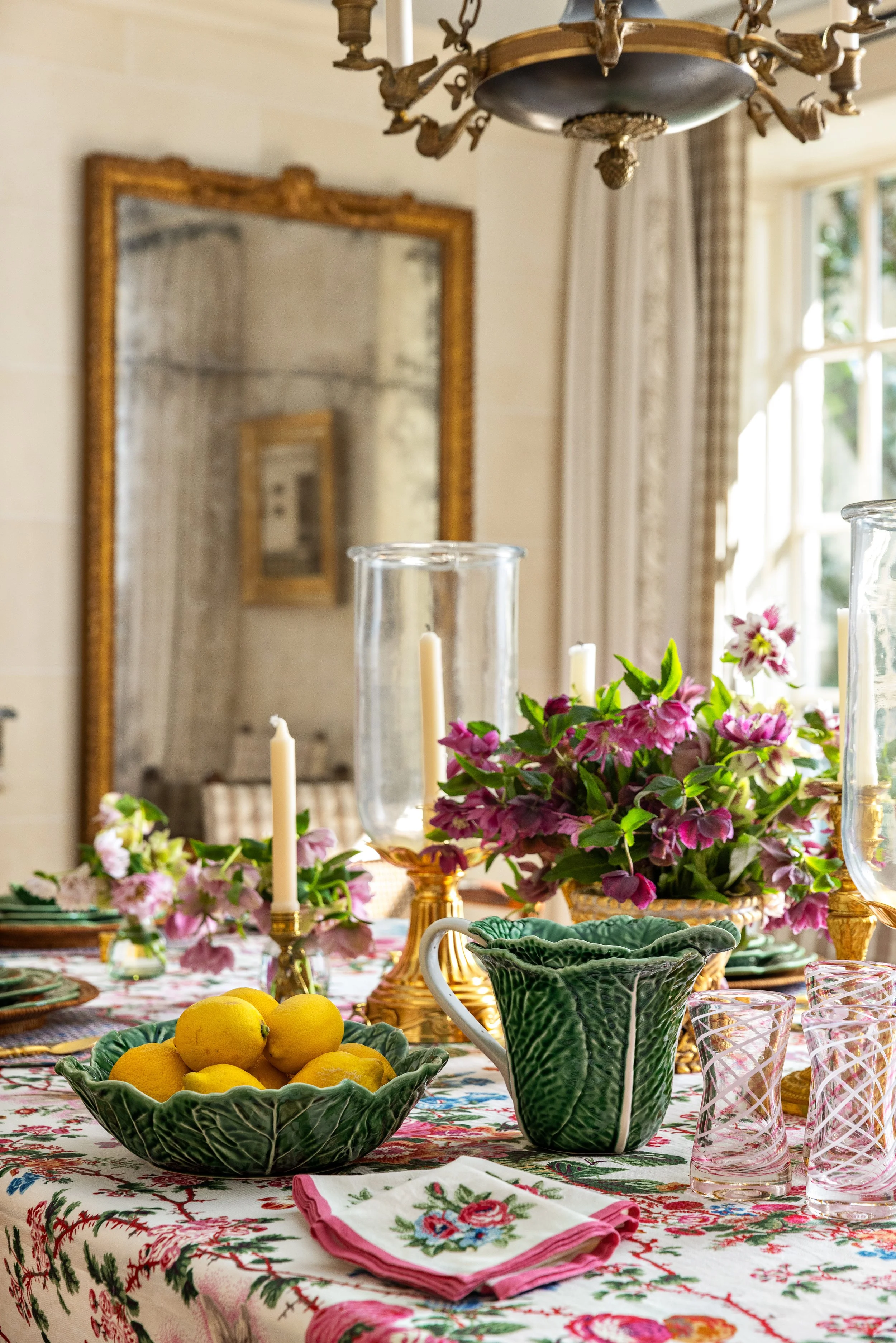 A table decorated for a festive gathering with a floral centerpiece, lemons, candles, and elegant glassware. The background features a large mirror and a window letting in natural light. By Kimberly Power