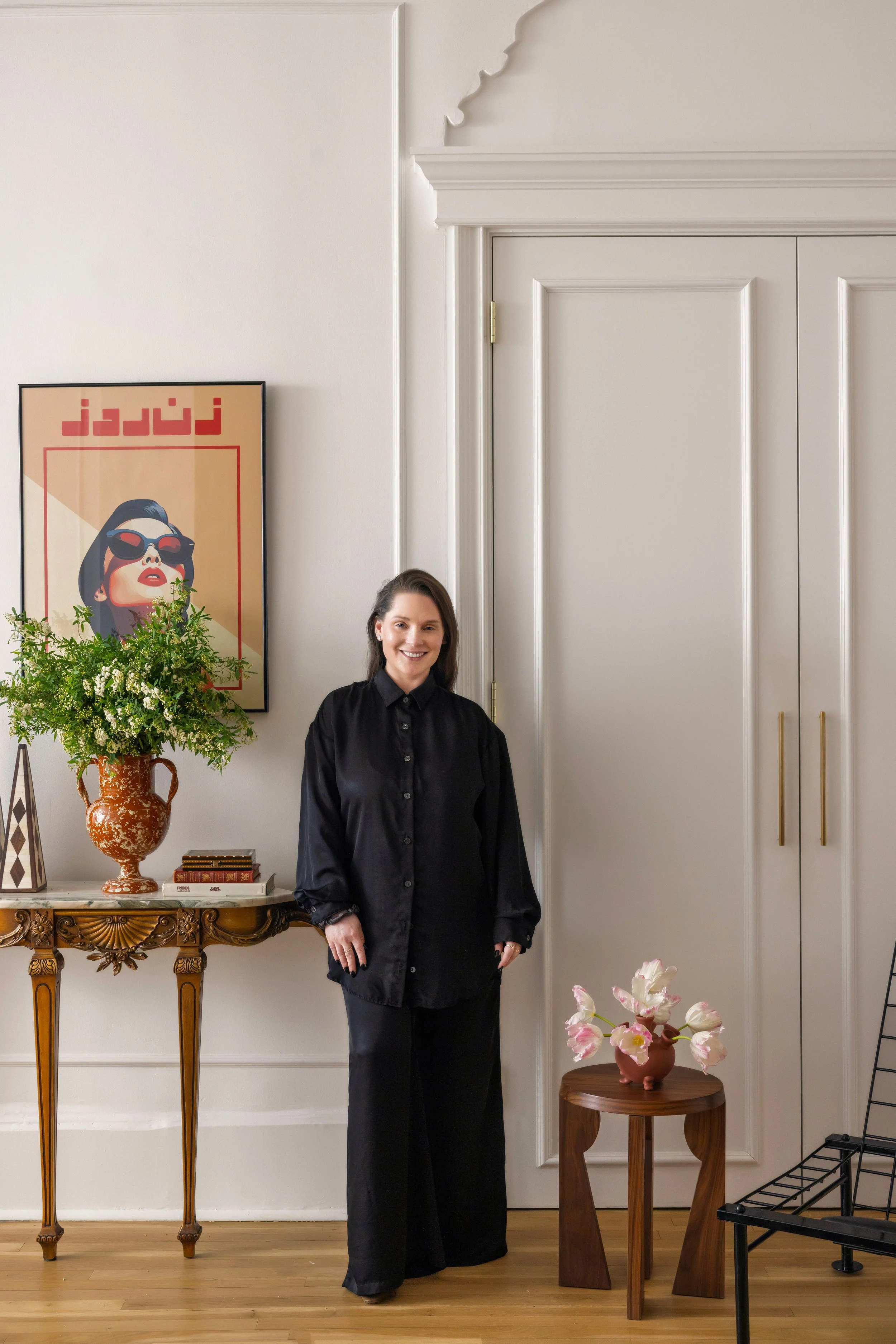 A woman with dark hair wearing a black outfit is standing in a room with decorated furniture and artwork. To her left, there is a wall with a framed poster of a woman with dark hair, sunglasses, and red lipstick. A pot of white and green flowers is on a decorative wooden table beneath the poster. To her right, there is a small wooden table with pink and white flowers in a pottery vase. The floor is wooden, and the wall and door behind her are white with ornate moldings.