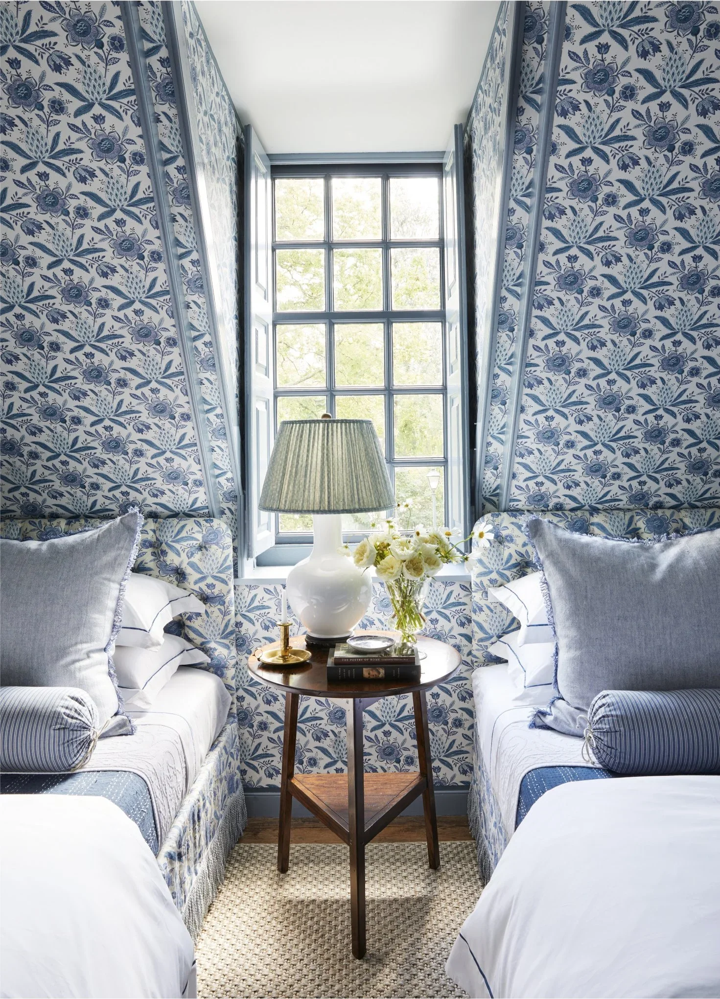 A cozy bedroom with two white beds and blue floral wallpaper. A small wooden table holds a white lamp, a glass vase with white flowers, and books. A window with open shutters lets in natural light.