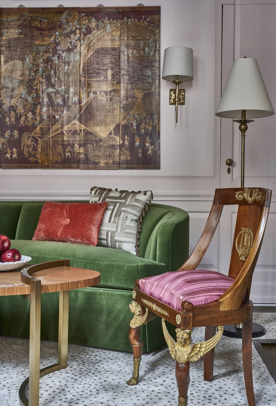 Interior of a living room with a green velvet sofa, decorative pillows, a wooden chair with pink upholstery, a small round wooden side table with gold legs, two wall lamps, a large tapestry with a historical scen. By Cody Damen Jenkins