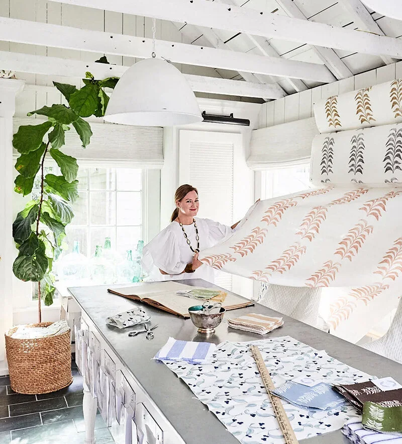 Heather Chadduck in a white top standing in a bright, white kitchen with a large table, plants, and patterned fabrics, holding up a piece of fabric.