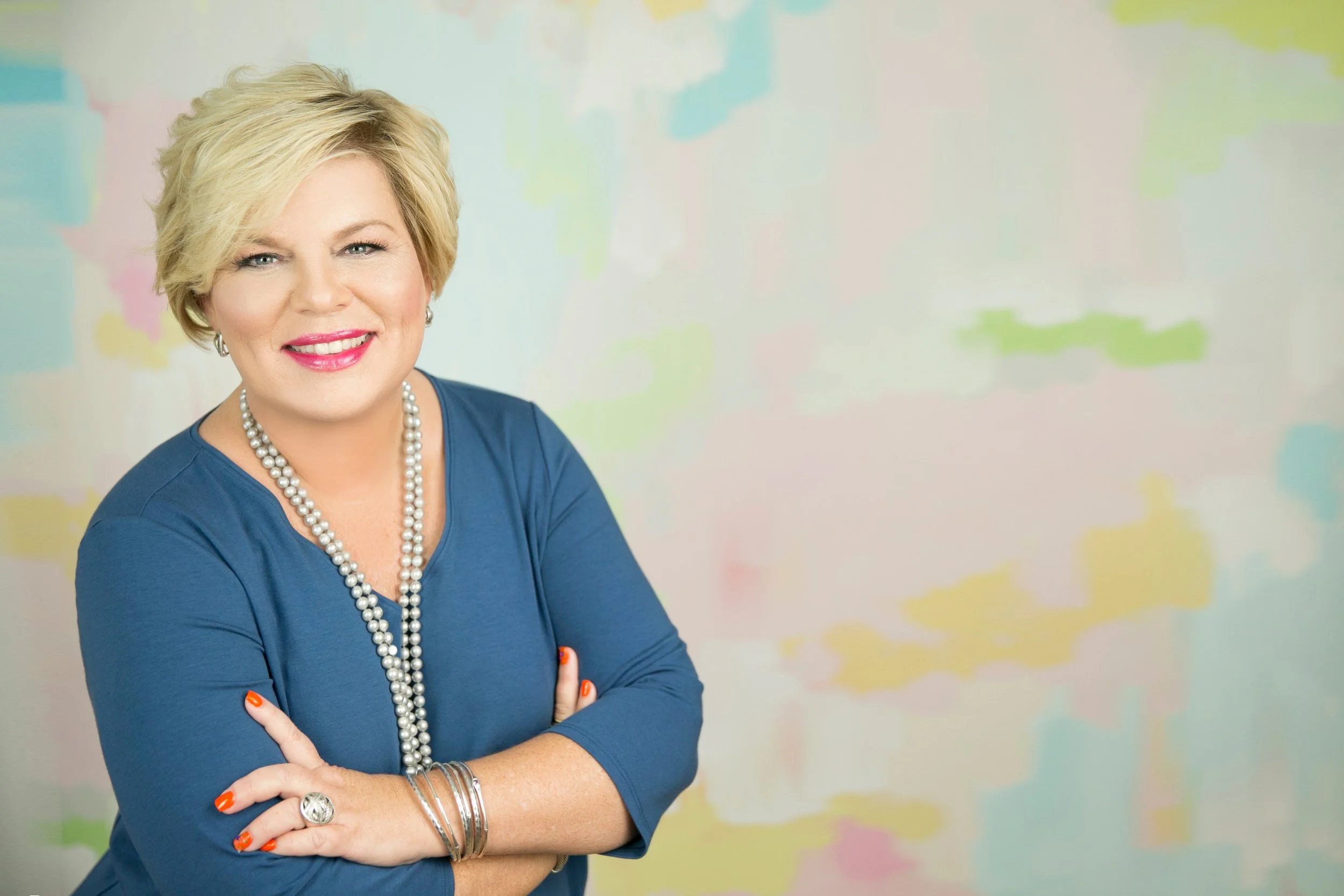 RJ Landin with short blonde hair, wearing a blue top, pearl necklace, and multiple bracelets, is smiling at the camera with her arms crossed in front of a colorful, pastel-patterned background.