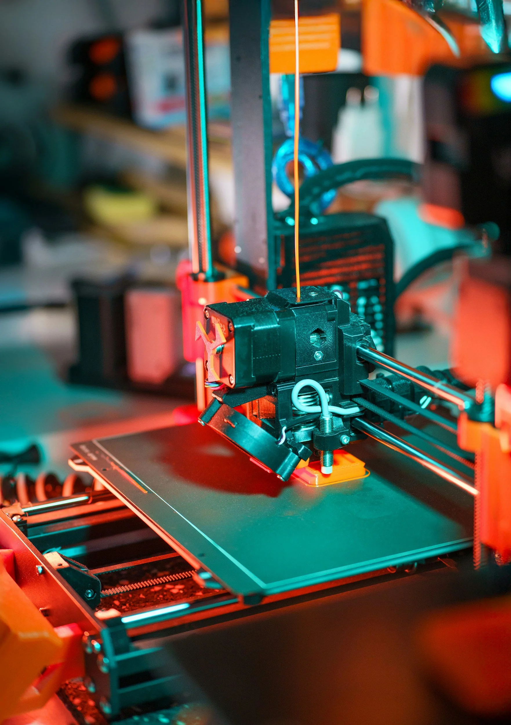 Close-up of a 3D printer with a nozzle extruding orange filament, illuminated by vibrant blue and red lighting.