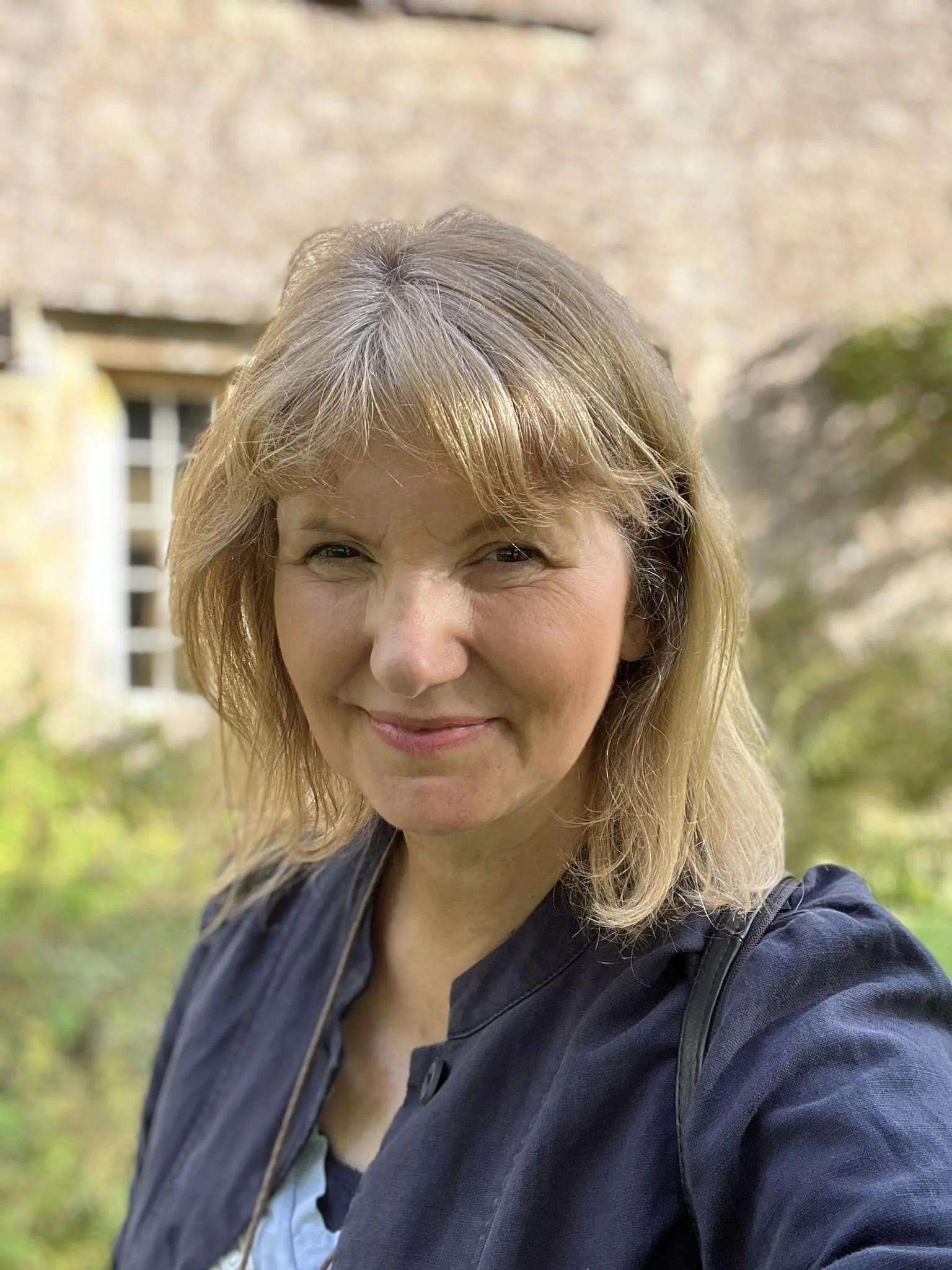 A woman with blonde hair smiling outdoors with a beige brick wall and some greenery in the background.
