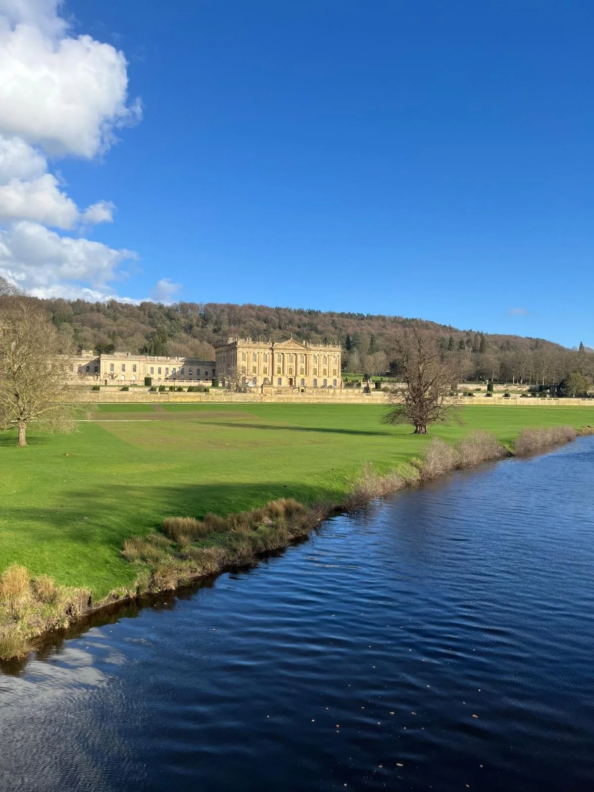 View of a grand historic building, likely a palace or estate, situated on a hill with a river in the foreground and green lawns and trees surrounding it under a clear blue sky.