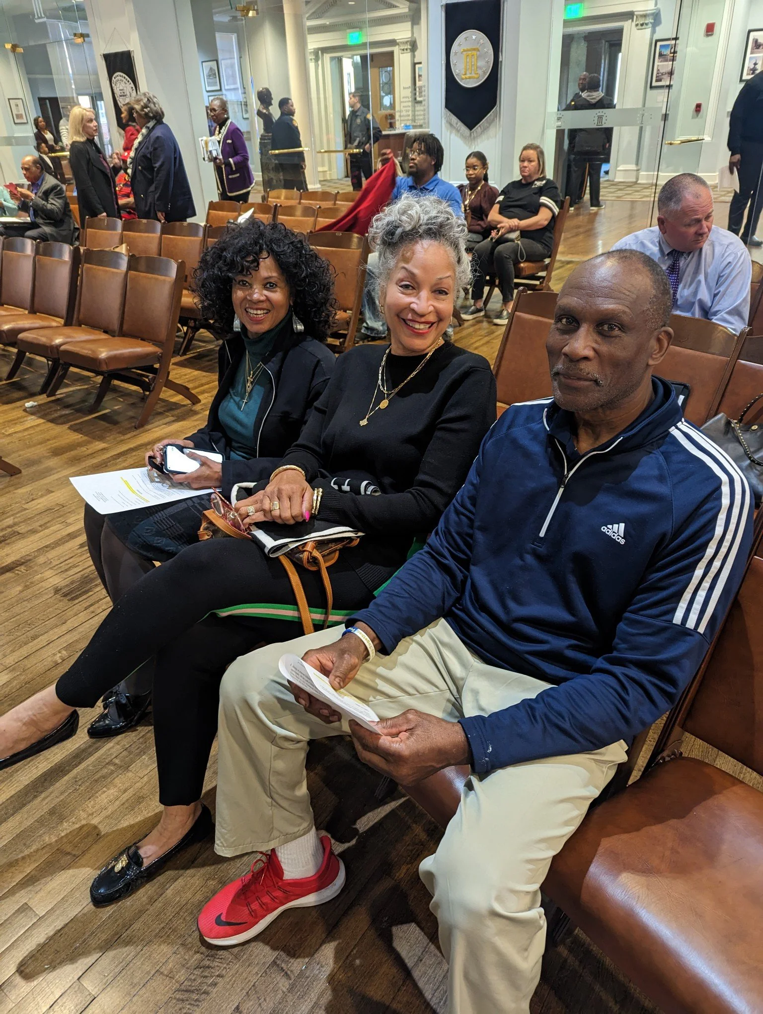 Three people sitting on chairs in a room with wooden floors and high ceilings, smiling at the camera. Behind them are more chairs and people sitting and walking around, with a glass wall, decorative banners, and framed pictures on the walls.