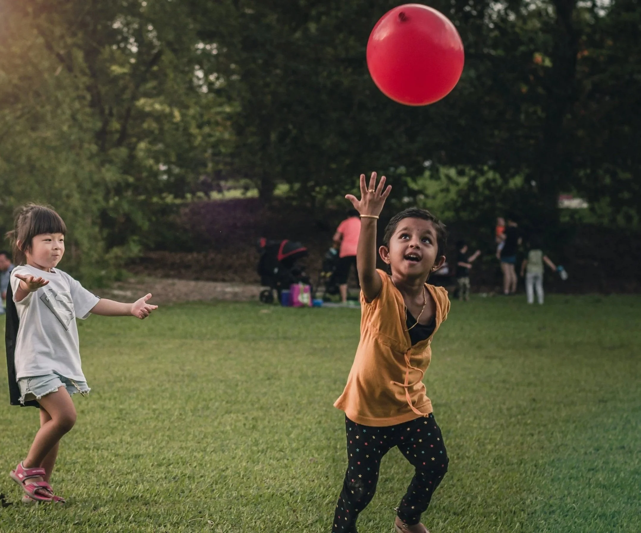 Two children playing with a red ball outdoors in a park with grass and trees.