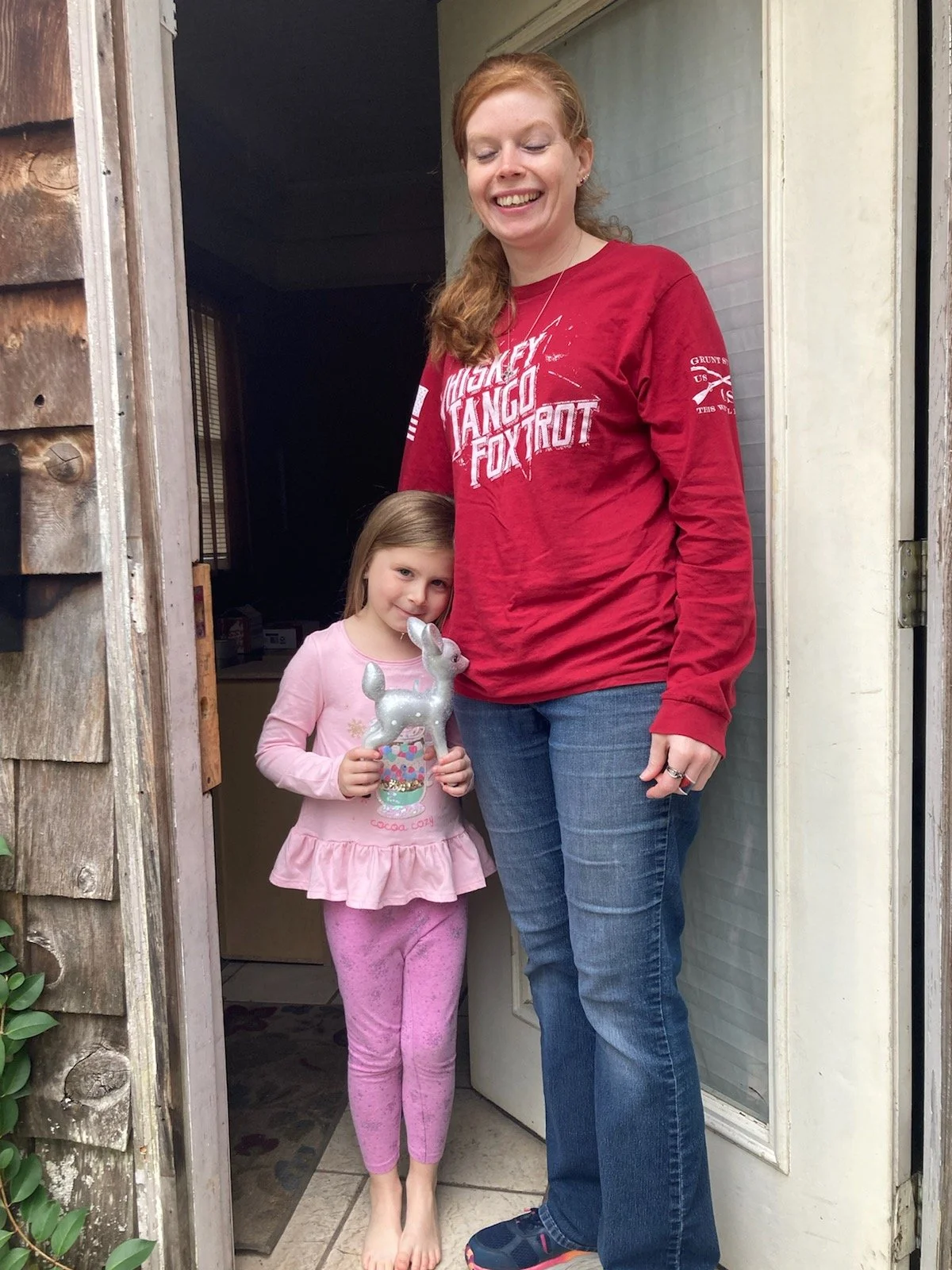 A woman and a young girl standing in a doorway. The woman is smiling and wearing a red long-sleeve shirt and jeans. The girl, holding a silver bunny-shaped balloon, is dressed in pink pajamas and barefoot. They are smiling together outdoors, near a wooden and white-siding structure.