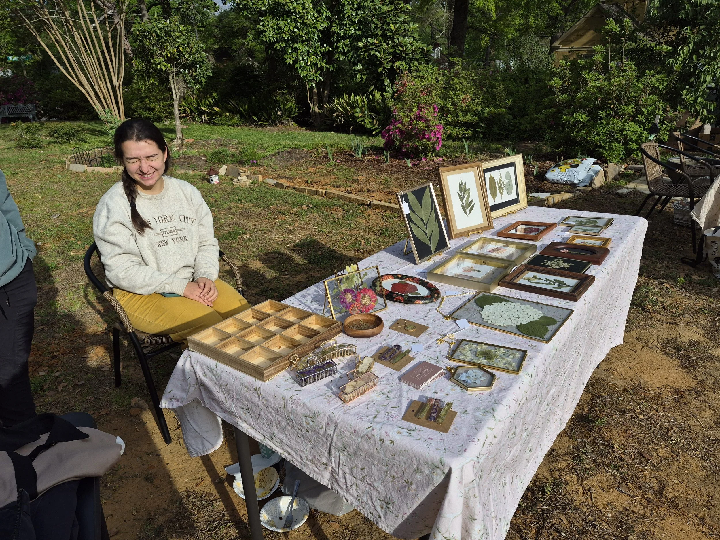 A woman sitting outdoors smiling with her eyes closed at a table displaying framed botanical artwork, jewelry, and small decorative items, with lush green trees and bushes in the background.