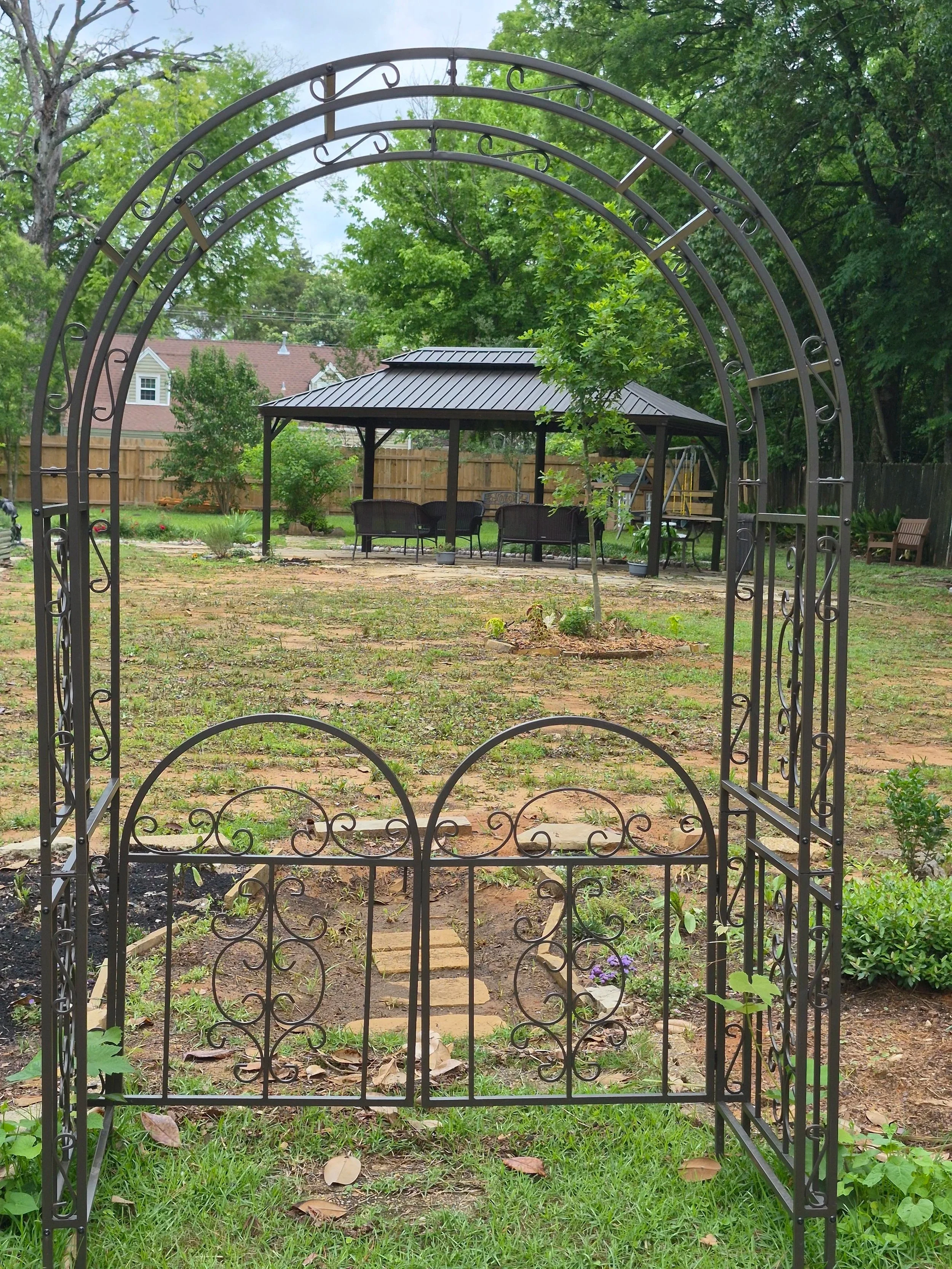View through an ornate metal garden gate into a backyard with a lawn, stepping stones, a gazebo with a table and chairs, trees, and a wooden fence.