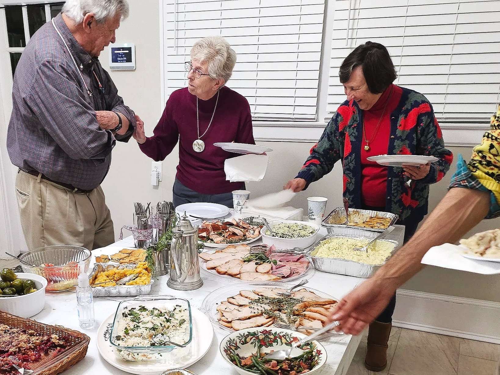 People serving and enjoying a holiday meal with various dishes including roasted meats, salads, and sides at a dinner table.