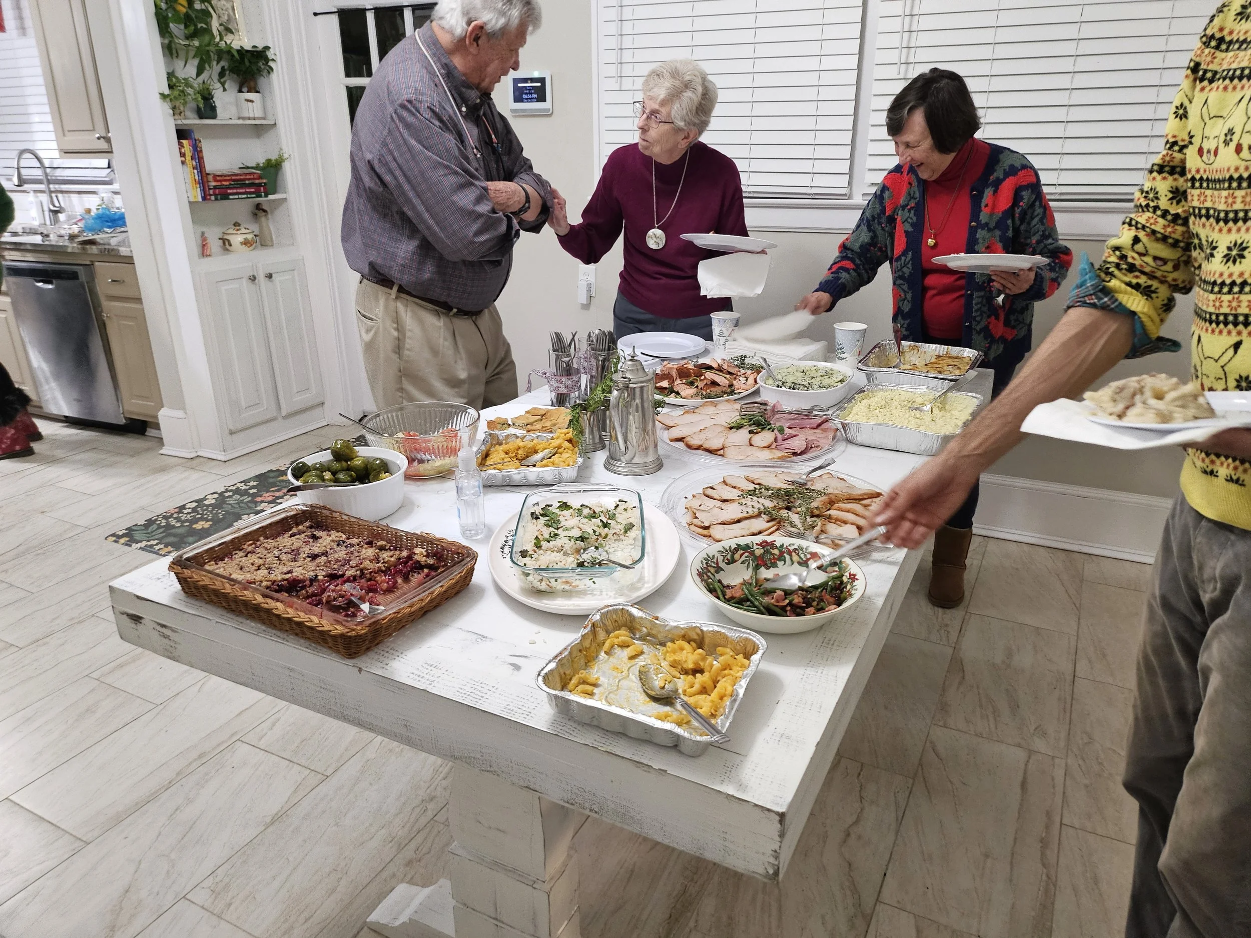 A group of elderly people gathered around a table with various dishes, including salads, meats, and casseroles, enjoying a meal together in a decorated kitchen or dining area.