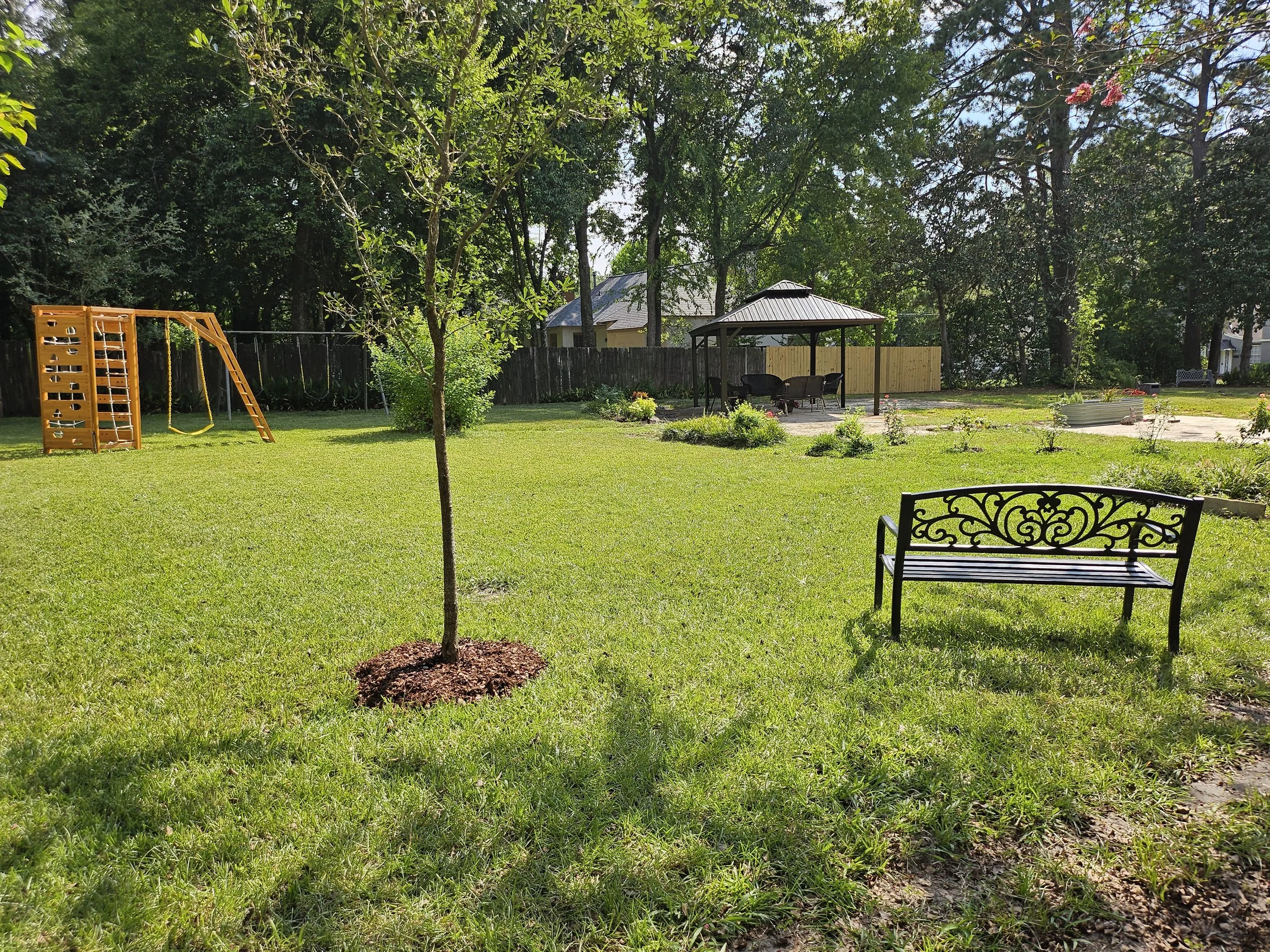 A green backyard with a small tree, a wooden playground set for children, a gazebo with outdoor seating, a flower bed, and a fenced yard surrounded by tall trees and other houses in the background.