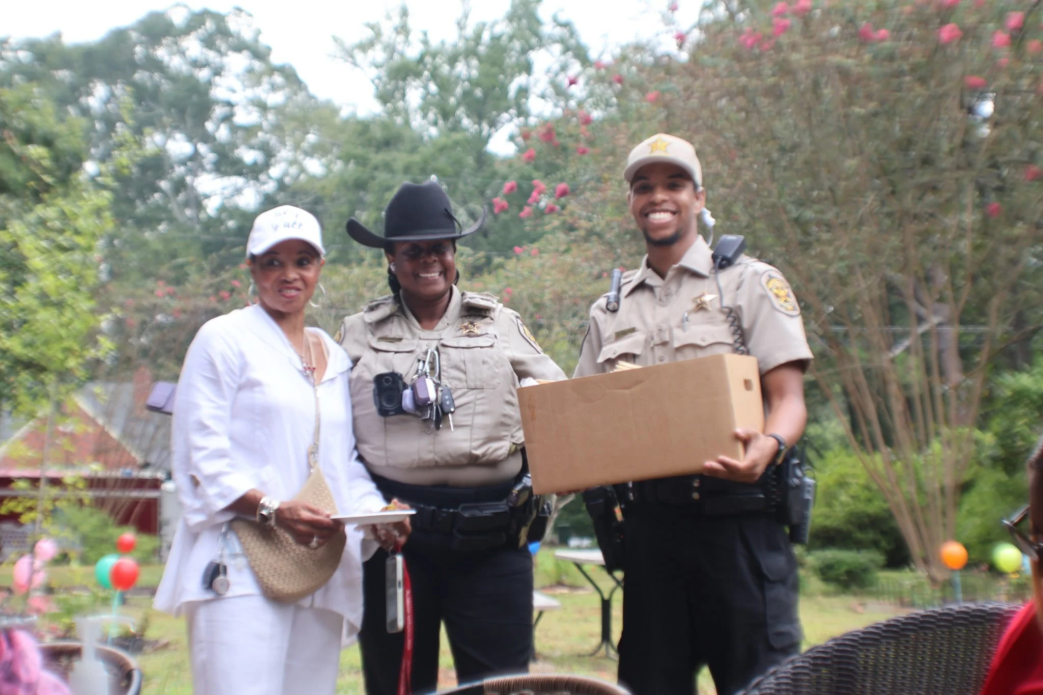 Three people, including two police officers in uniform and a woman in white, posing together outdoors with trees and pink flowers in the background. One officer is holding a cardboard box.