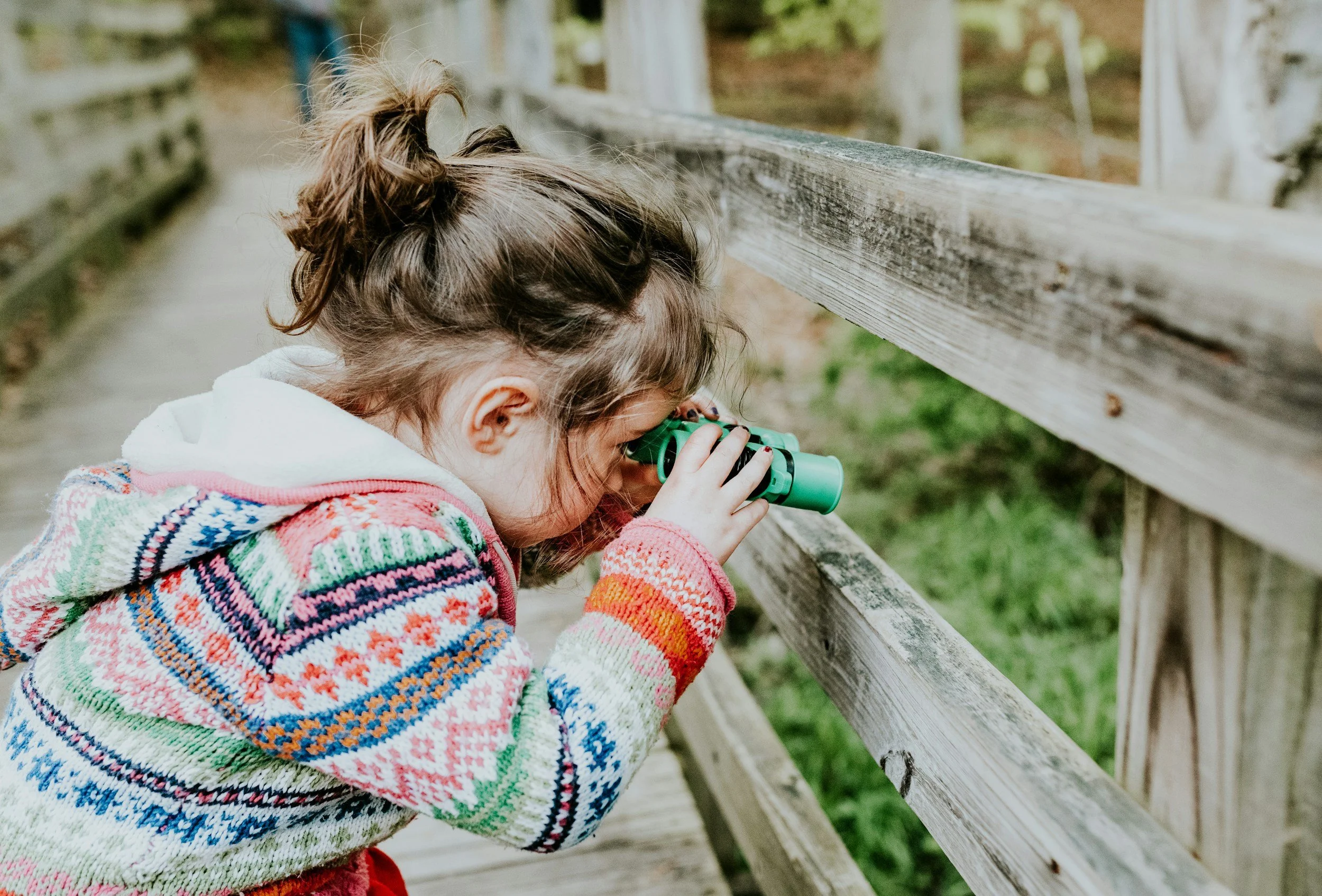 A young girl with brown hair in a messybun looking through green binoculars, wearing a colorful knitted sweater, leaning against a wooden fence outdoors.