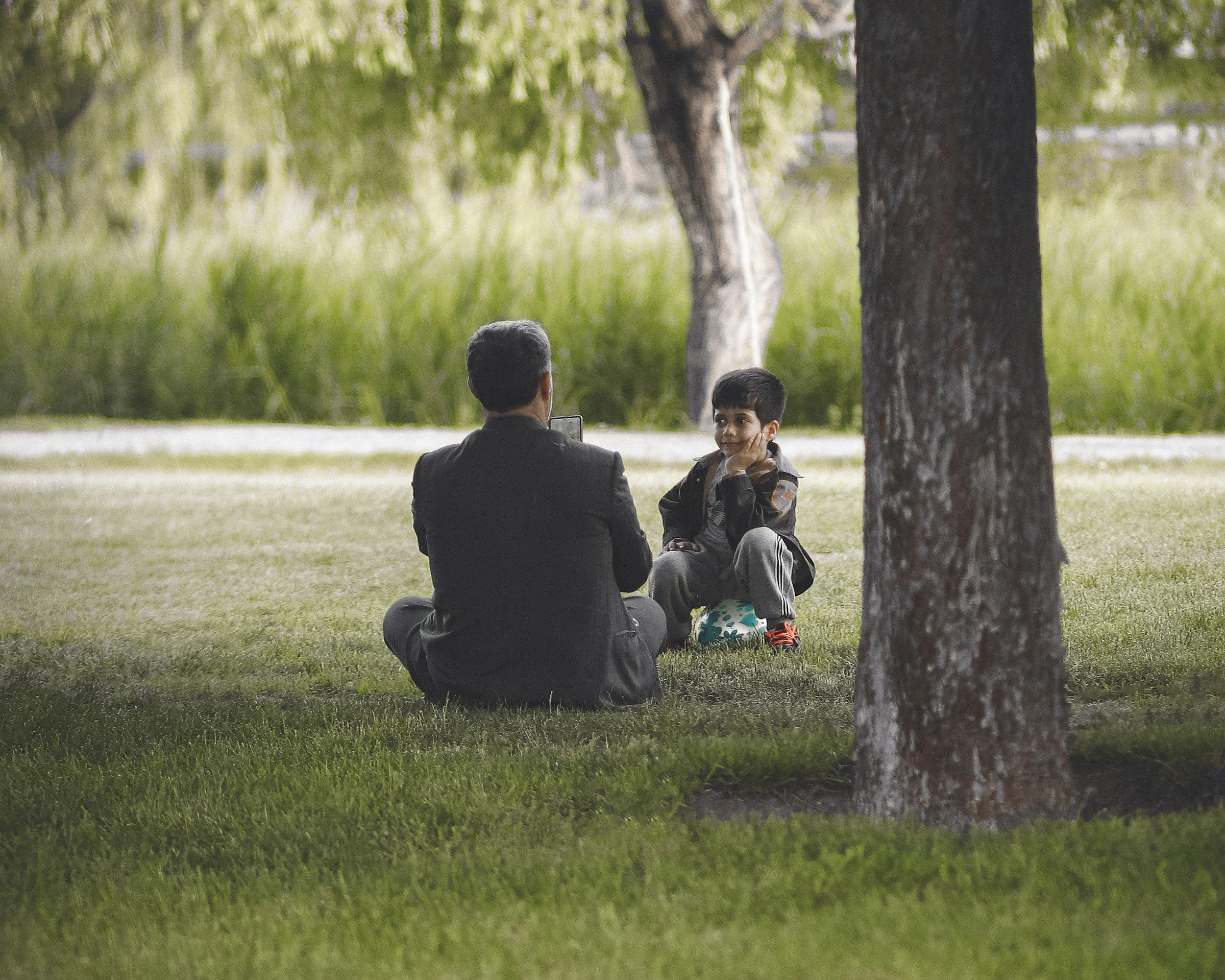 A man and a boy sitting outdoors on grass near a tree in a park, with the boy resting his chin on his hand and looking at the man.
