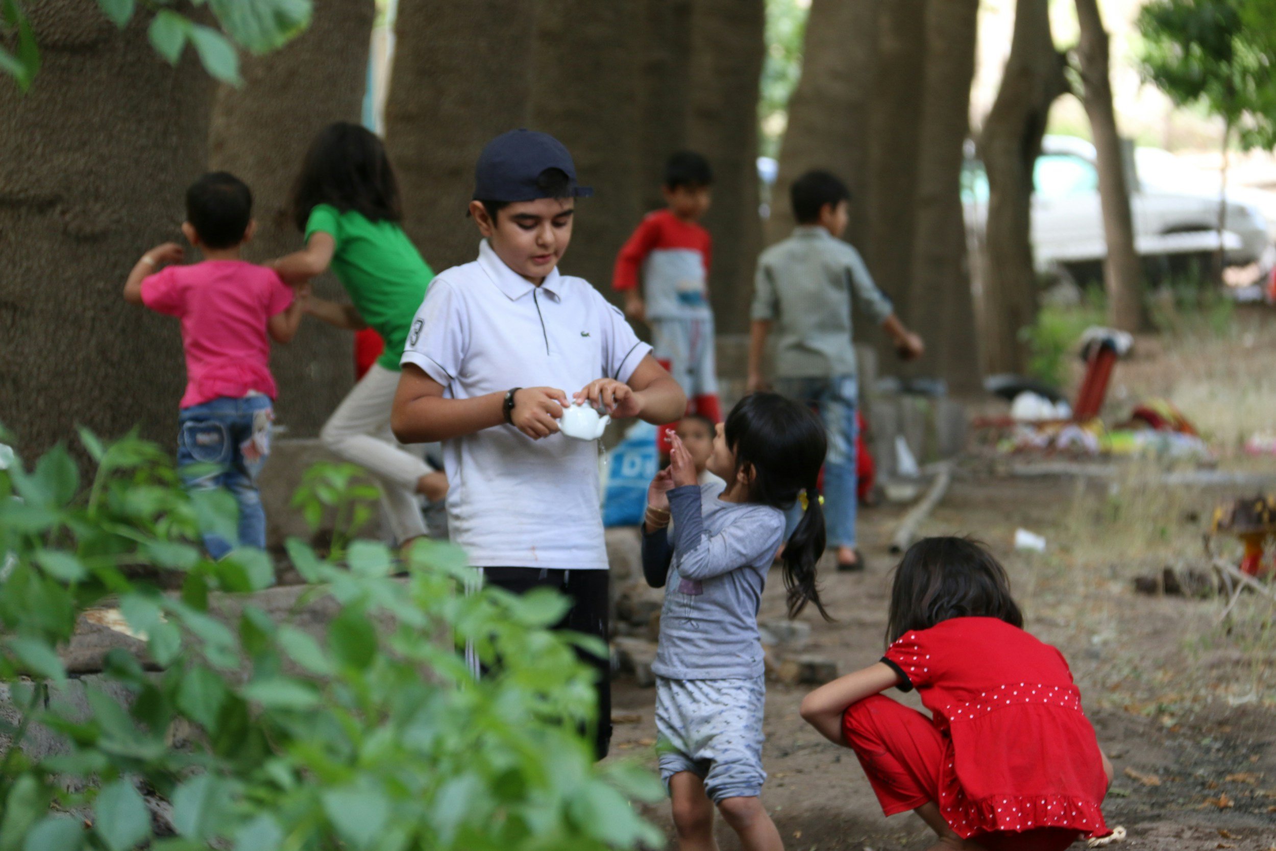 Children playing outdoors in a park, some standing and some sitting, with trees and green foliage in the background.