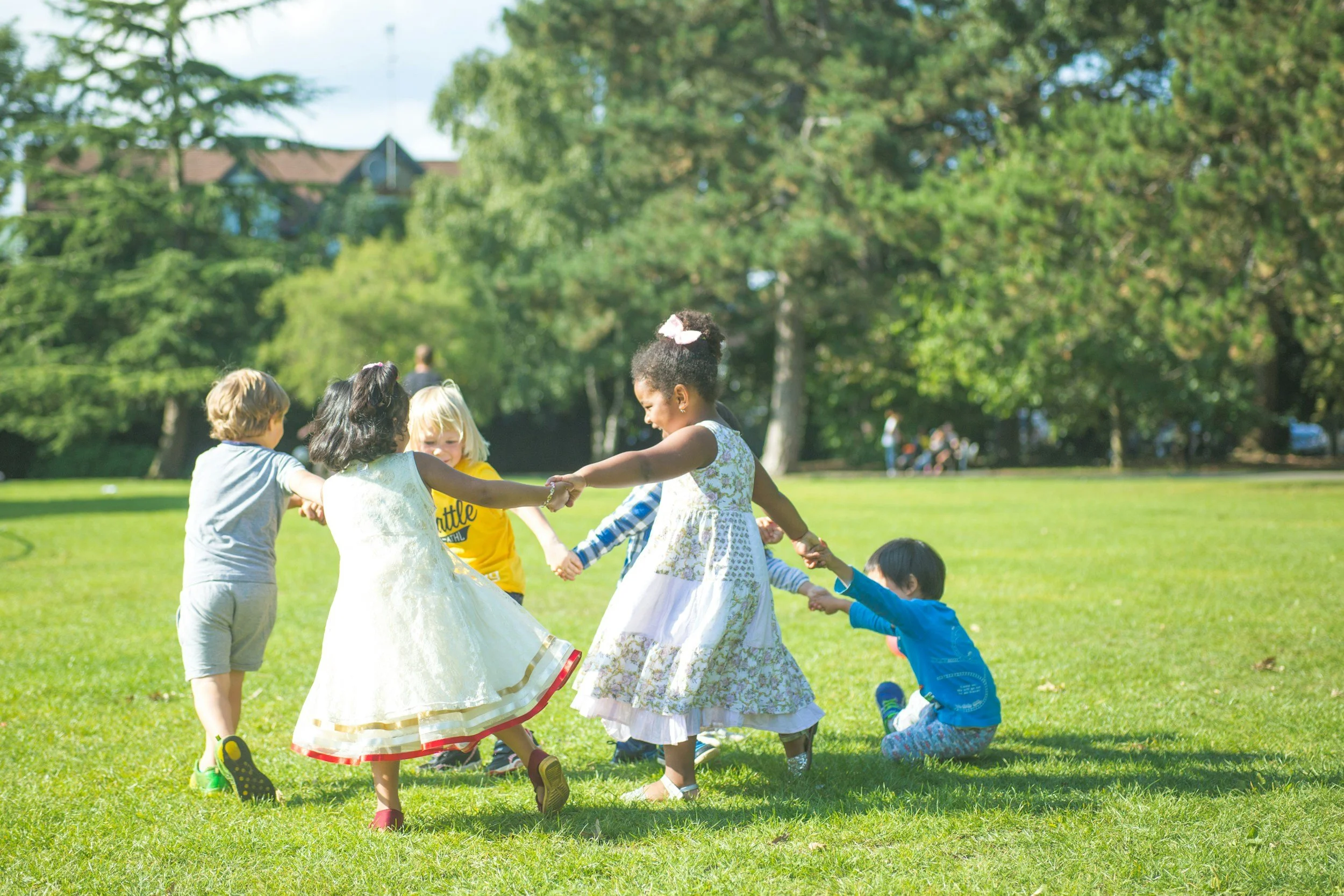 Kids holding hands and playing in a circle on a sunny grassy field in a park with trees and houses in the background.