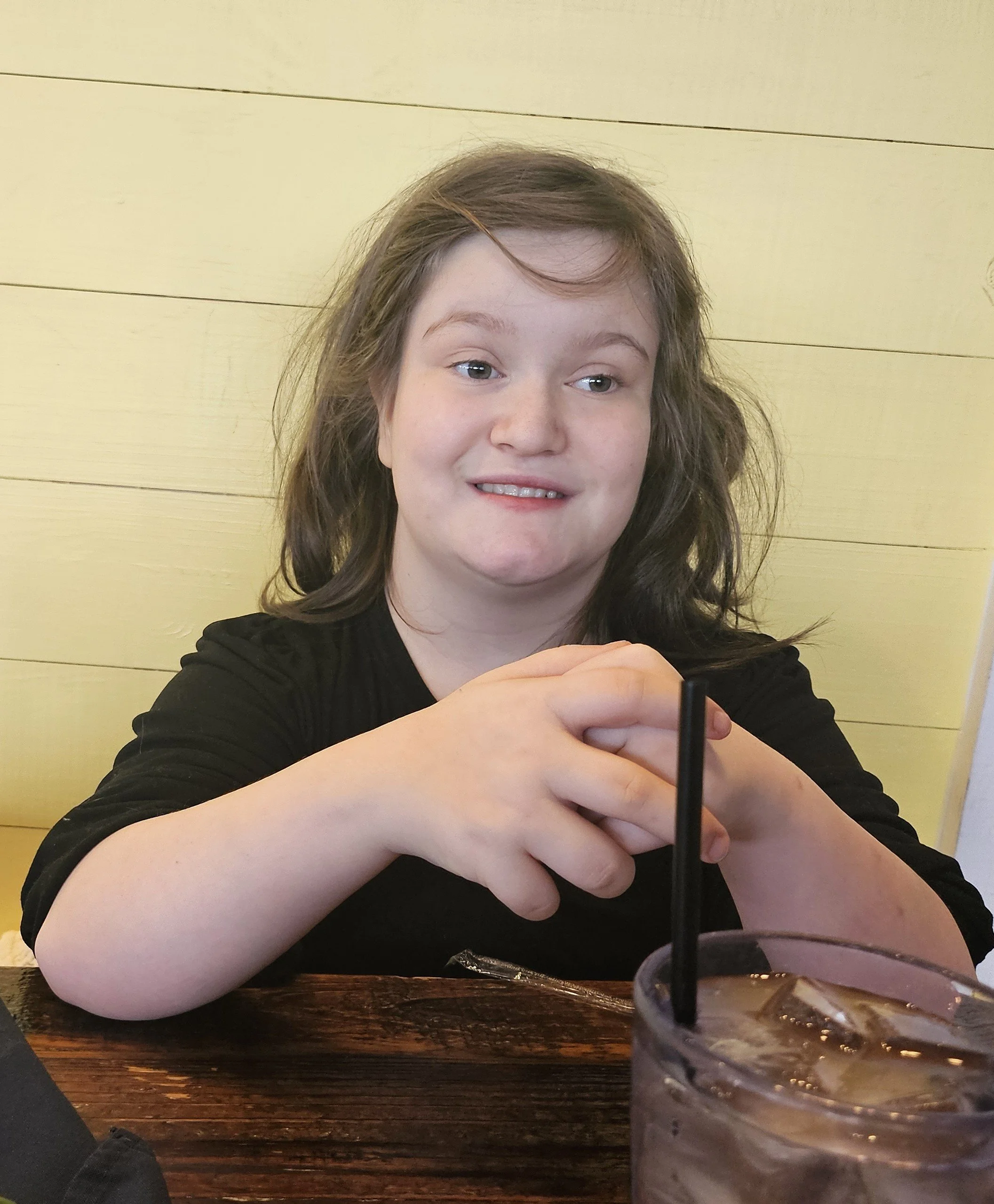 A woman with brown hair and a black shirt sitting at a wooden table, smiling slightly, with a drink in a clear cup with ice and a black straw in the foreground.