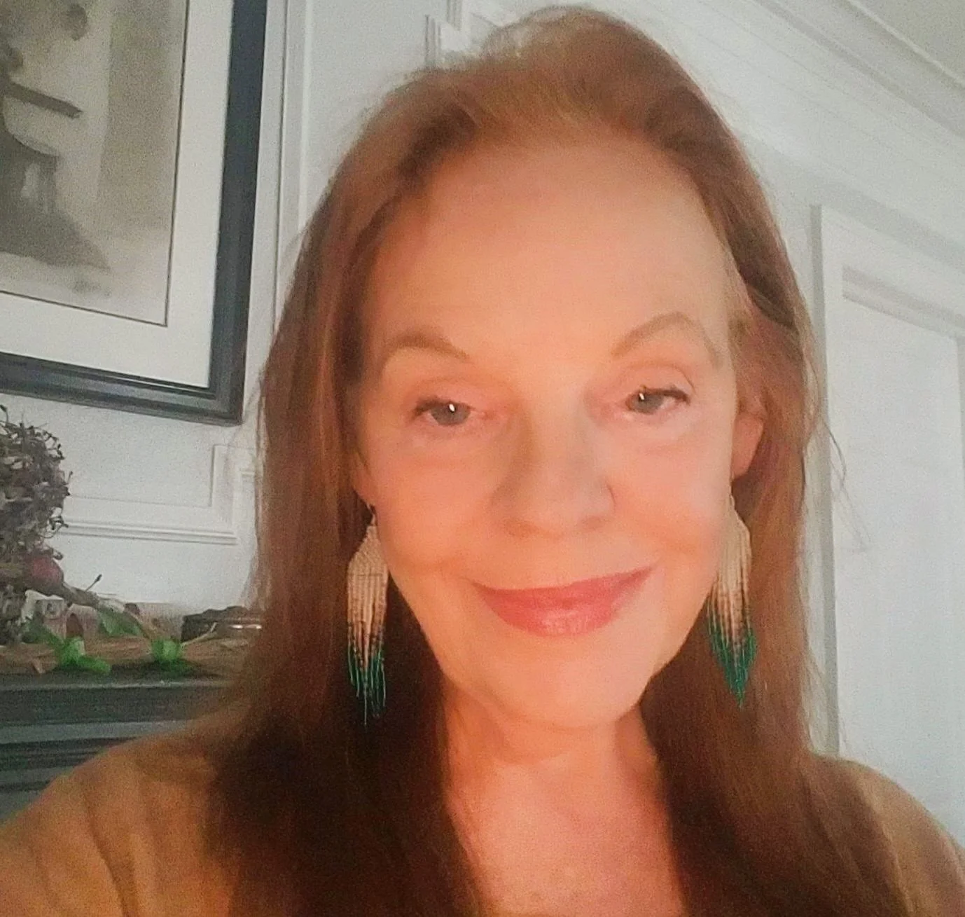 A woman with long auburn hair and light makeup, smiling softly, wearing colorful earrings, in an indoor setting with white walls and a framed picture in the background.