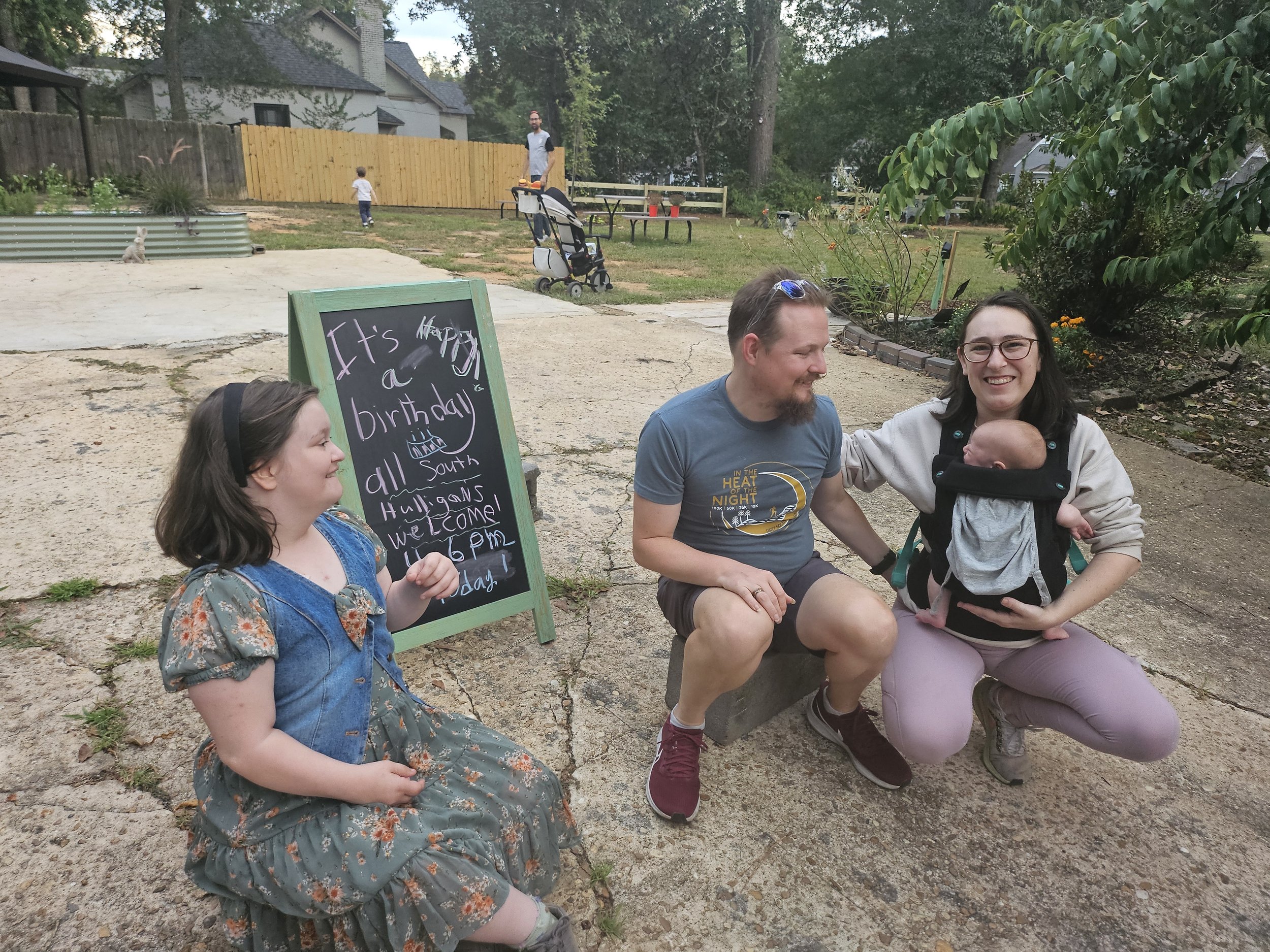 A group of people sitting on a park bench, smiling and enjoying a conversation. There is a child and a man in a blue shirt, and a woman holding a baby carrier. A chalkboard sign with handwritten birthday wishes is in front. In the background, a man and child are near a wooden fence, with trees and houses beyond.