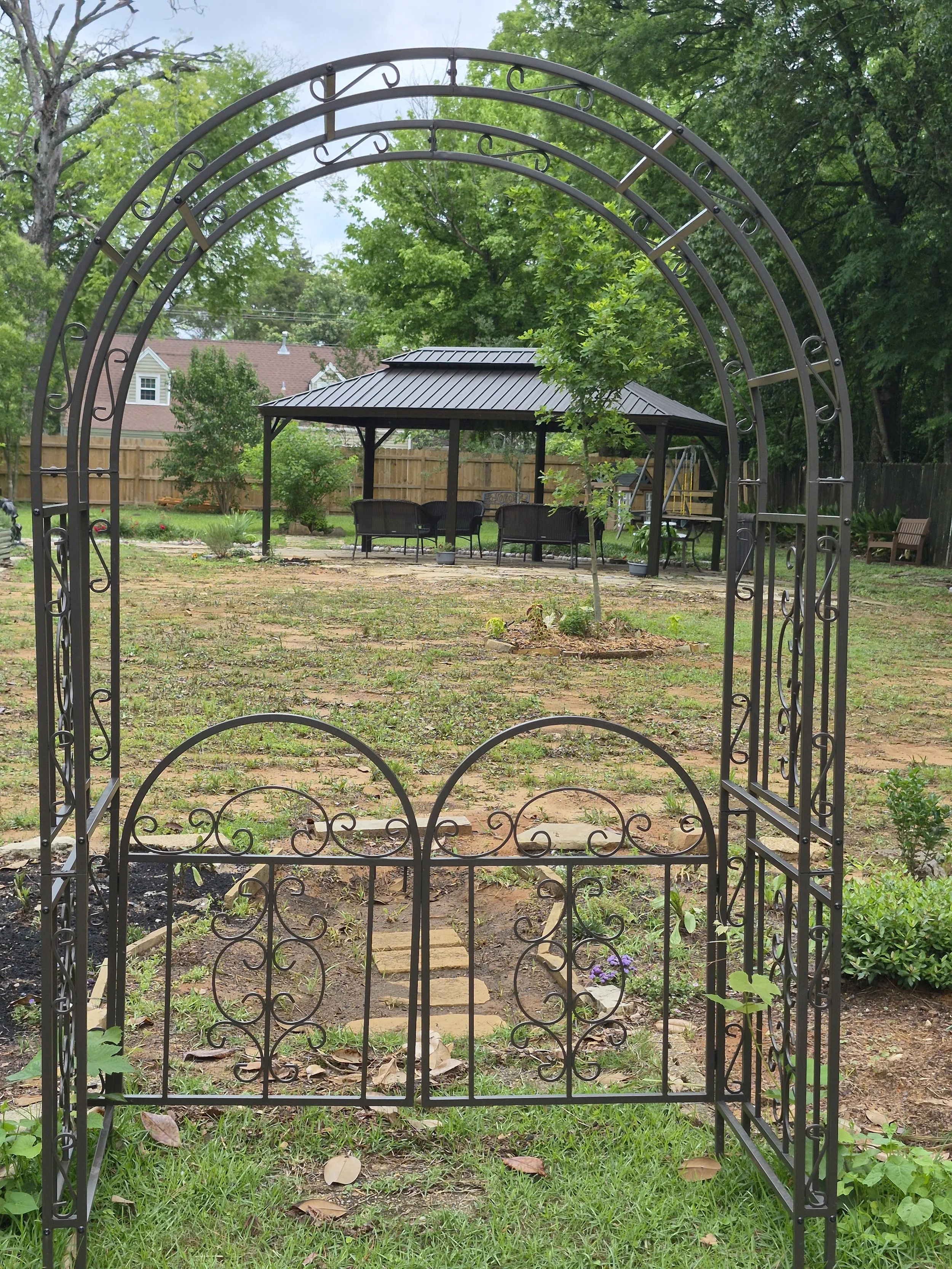 View of a backyard with a black metal garden archway in the foreground, leading to a patio area with a gazebo, outdoor furniture, and surrounding greenery including trees and bushes, with a wooden fence in the background.