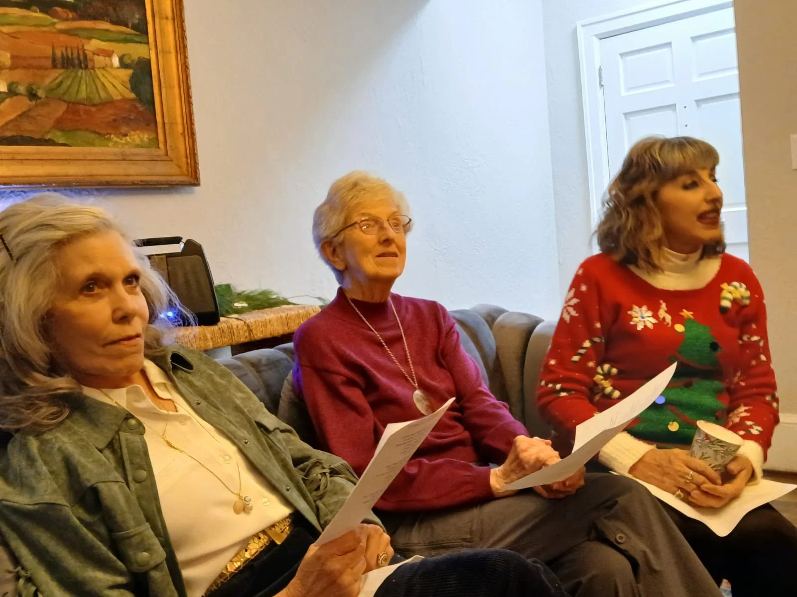 Three women sitting on a couch at an indoor gathering, holding papers, with a Christmas-themed sweater and holiday decorations in the background.